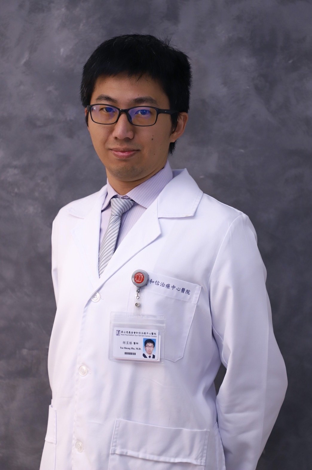 A male doctor in a white coat with a name tag, standing against a gray background.