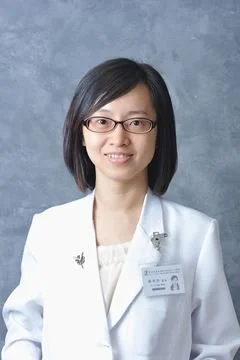 A woman with glasses and shoulder-length black hair wearing a white coat and a name badge stands against a blue-gray background.
