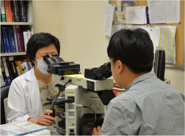 A woman scientist wearing a face mask examining a sample through a microscope in a laboratory, with a man colleague observing, amidst shelves with binders and papers in the background.