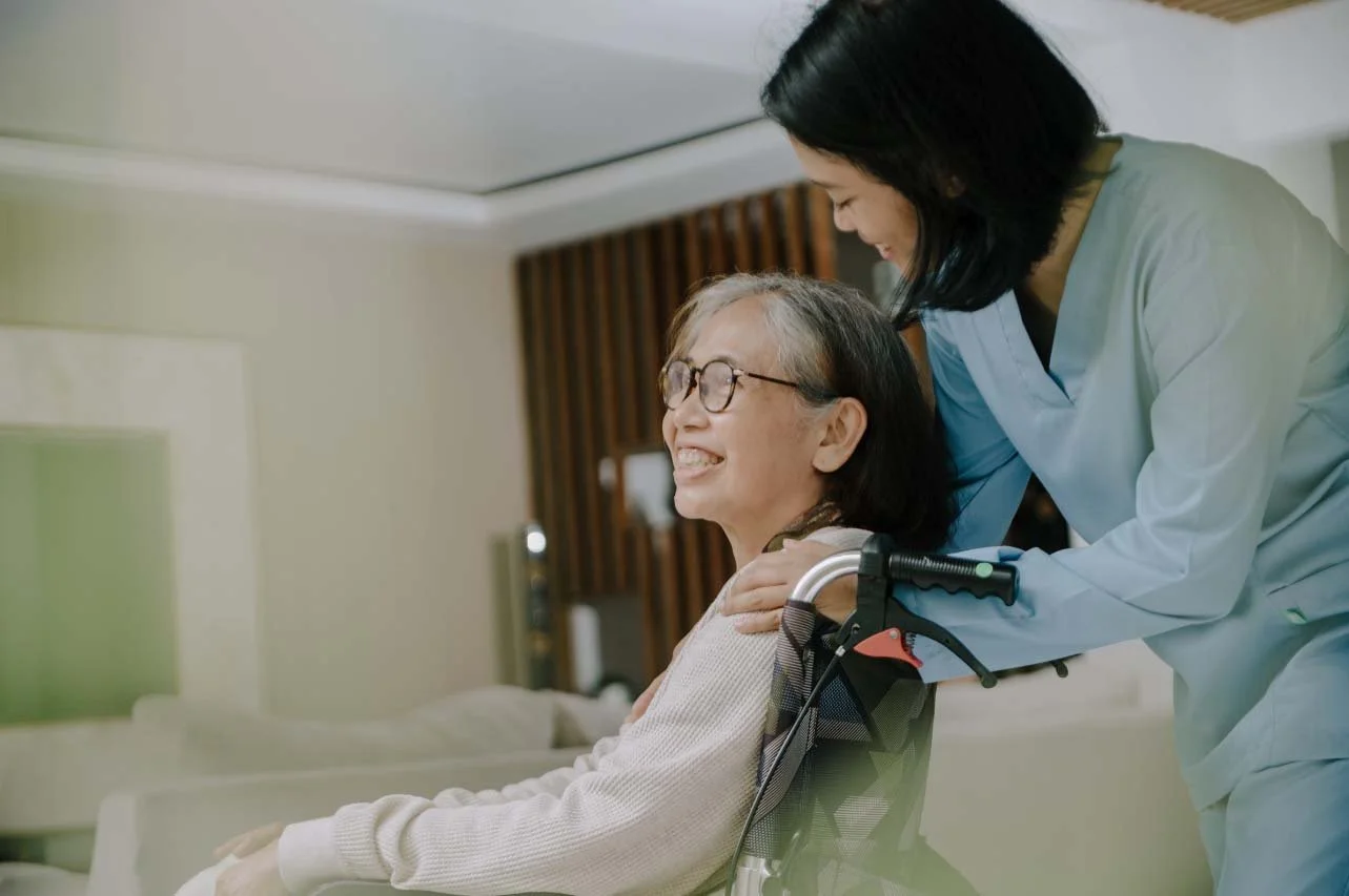 A healthcare worker helping an elderly woman in a wheelchair, smiling and showing care. The woman has glasses and is in a medical or home care setting.