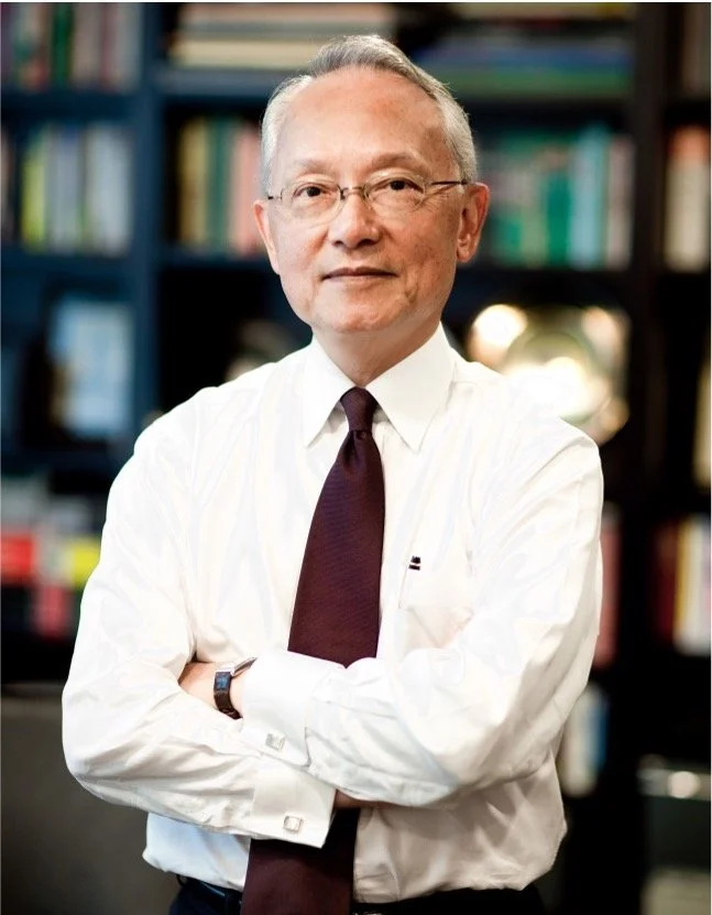 An older man with glasses, wearing a white dress shirt and a dark tie, standing with arms crossed in a room with bookshelves filled with books in the background.