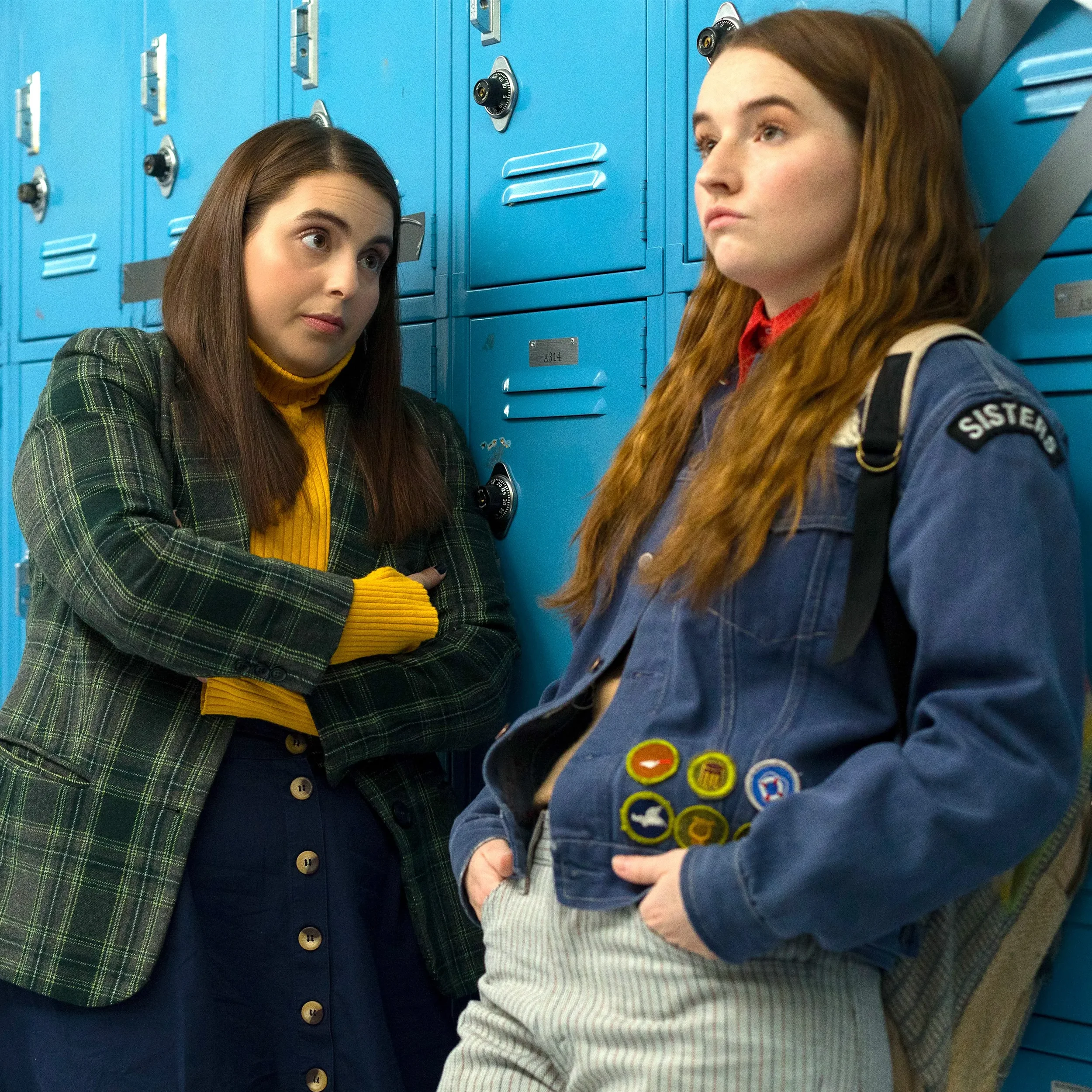 Molly and Amy from the film Booksmart standing next to lockers in highschool. Molly is looking at Amy and Amy is looking away.
