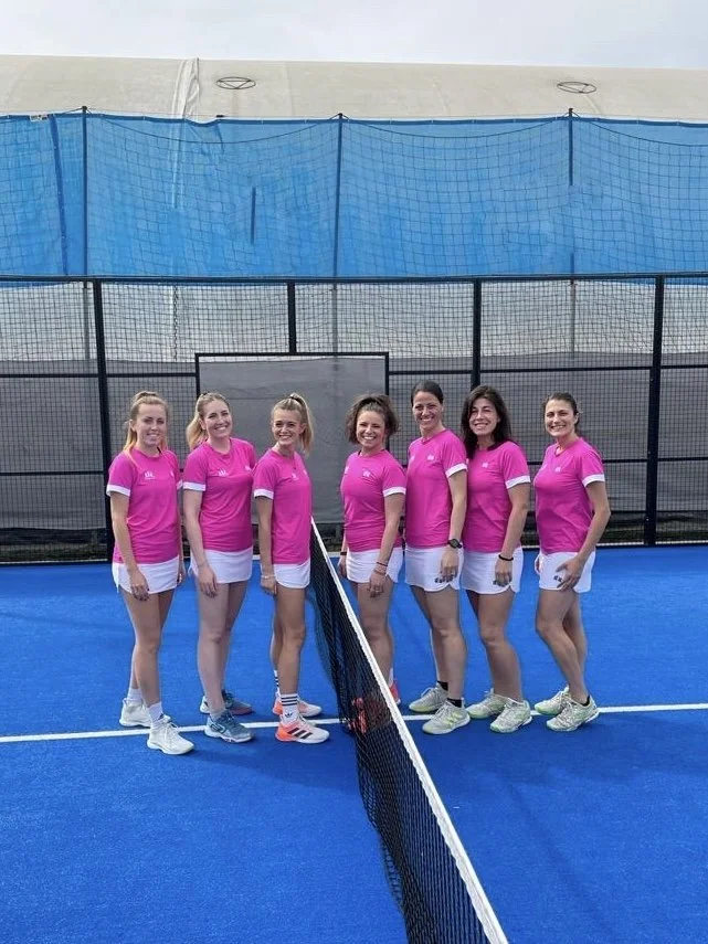 Sette donne su un campo da tennis indoor indossando magliette rosa e pantaloncini bianchi, posano dietro la rete.