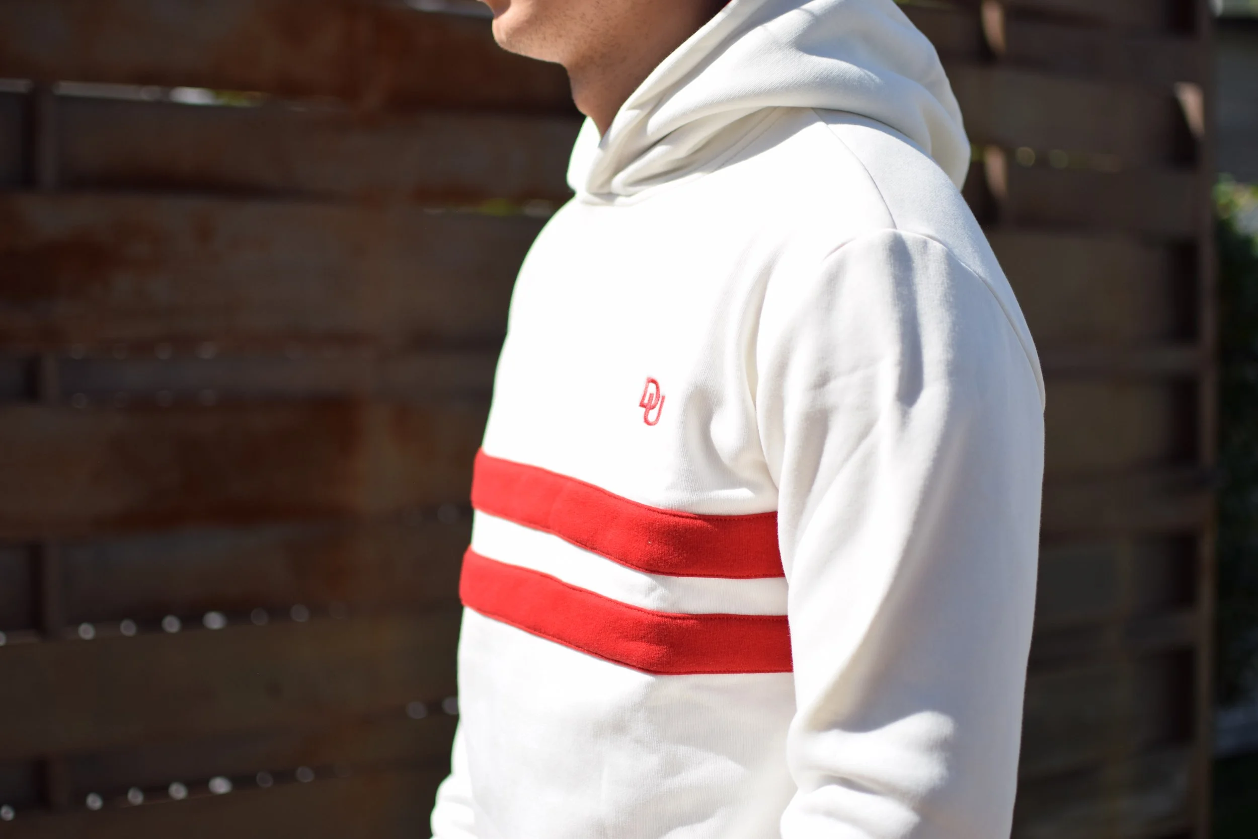 Persona indossando felpa bianca con bande rosse orizzontali e cappuccio, sfondo di recinto in legno.
