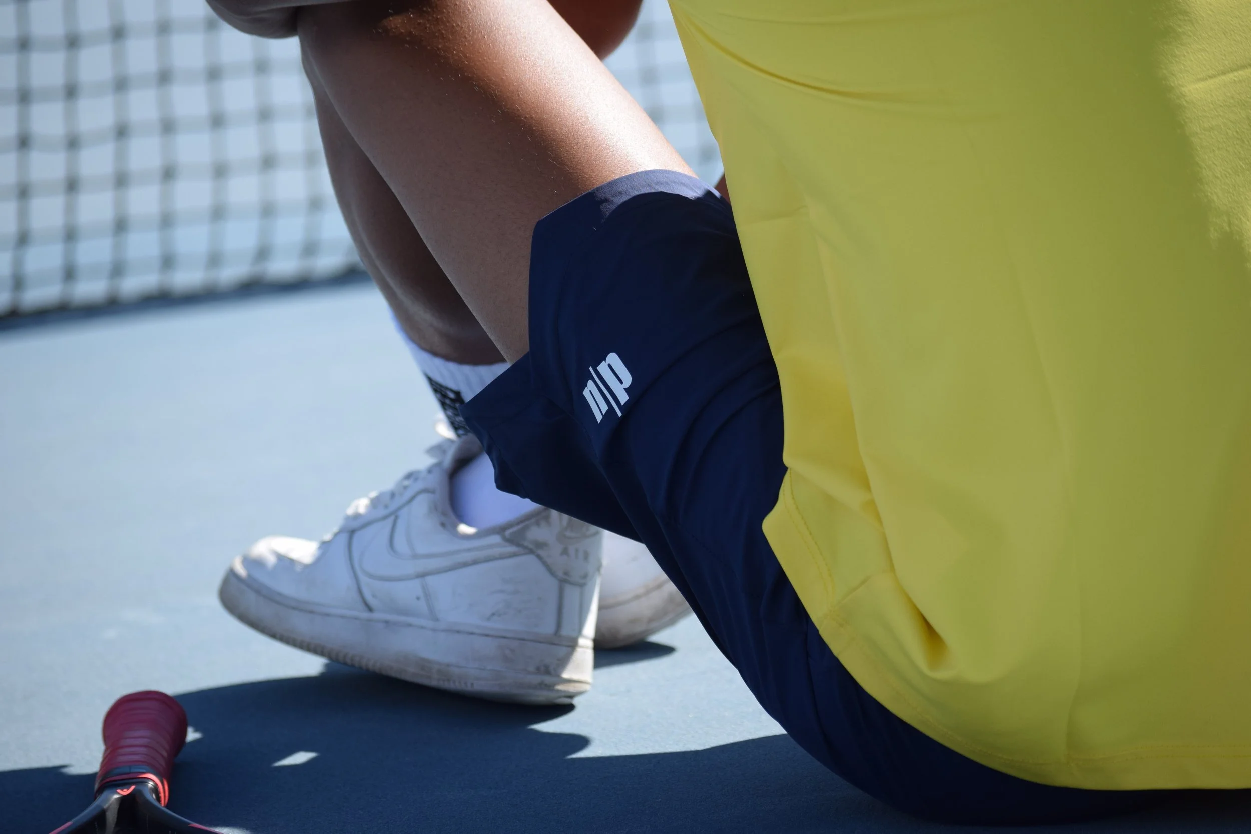 Gamba e scarpe da tennis di un giocatore di tennis su un campo da tennis, con rete visibile in sfondo.
