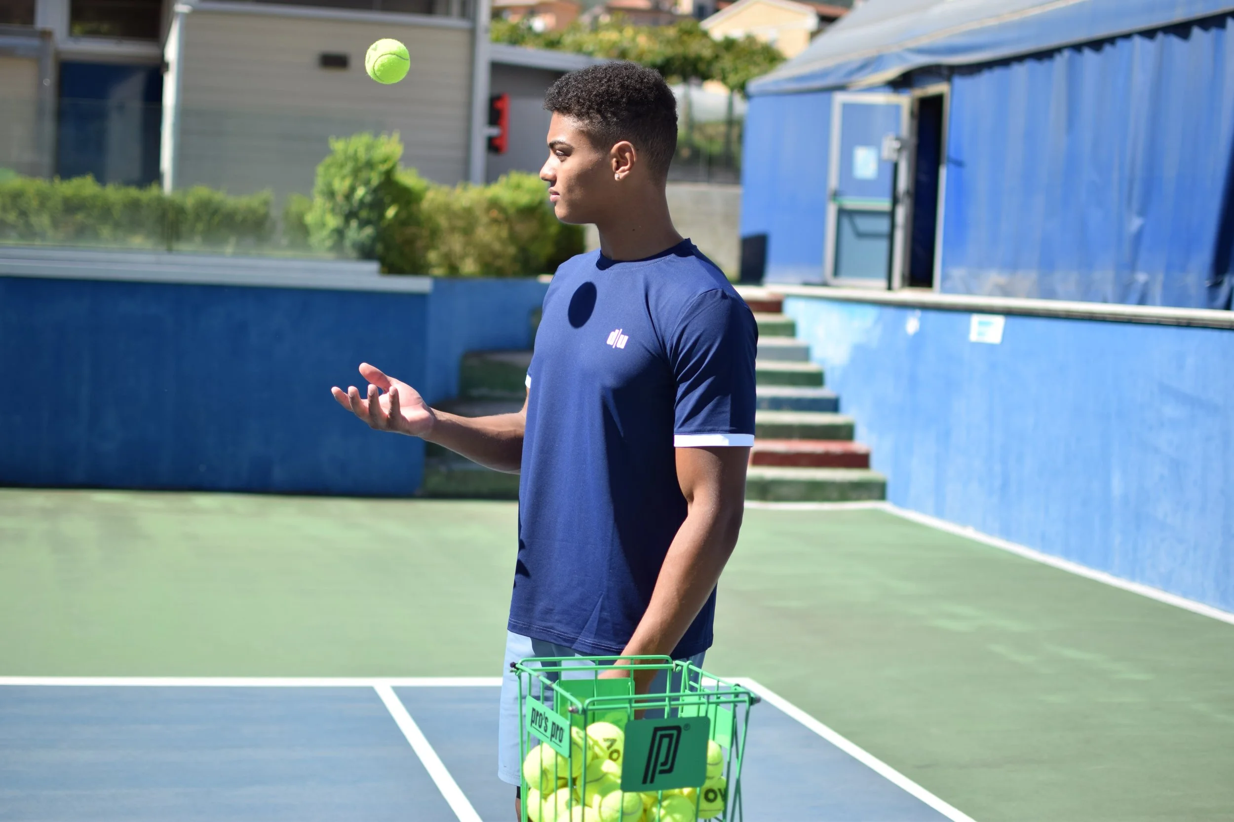 Giovane in piedi su un campo da tennis con una cesta di palline da tennis, mentre fa cadere una pallina in aria con una mano, in un ambiente all'aperto.