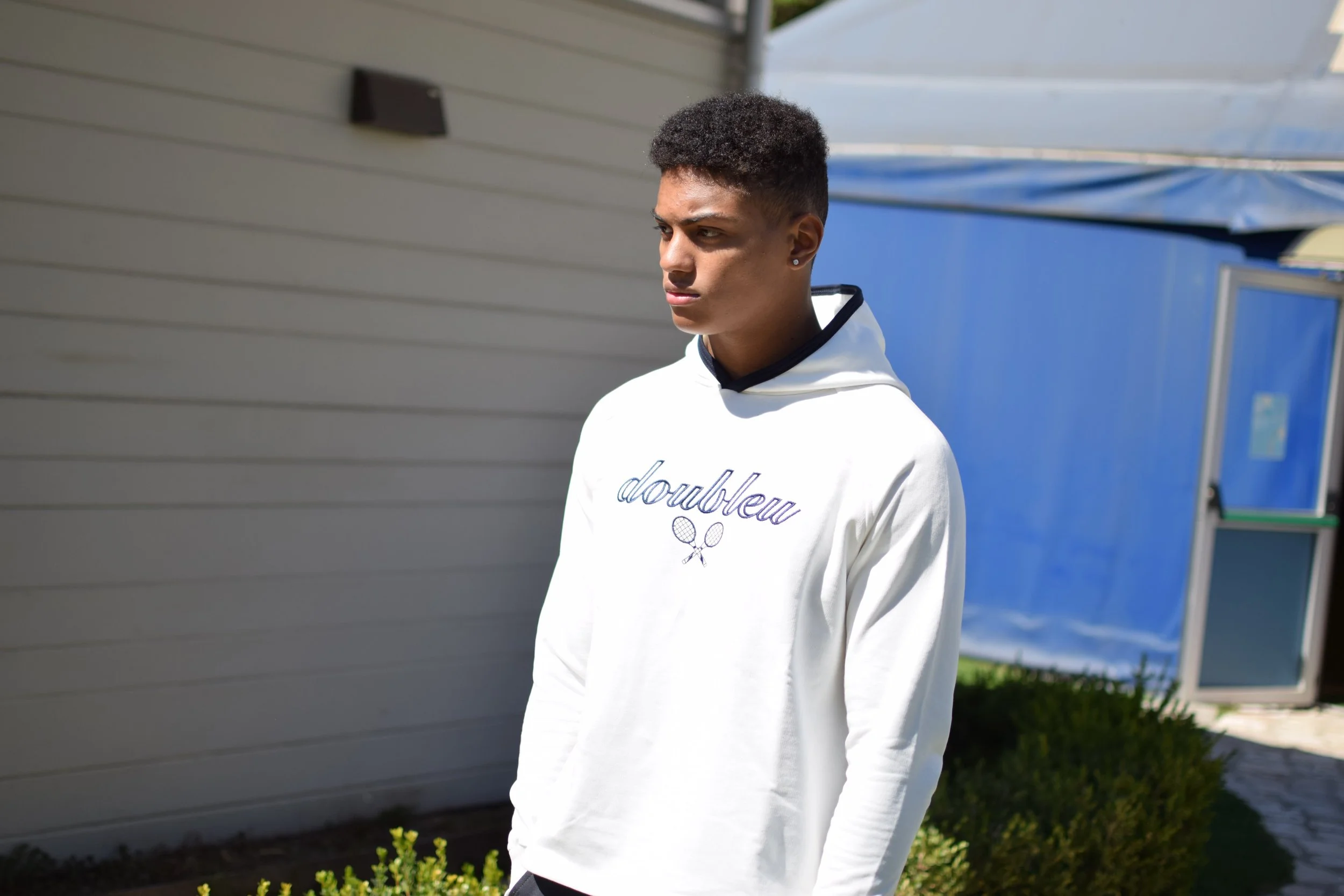 Ragazzo giovane che indossa felpa bianca con scritta "doubleu" e disegni di racchette da tennis, si trova all'aperto vicino a una parete beige e a una struttura blu.