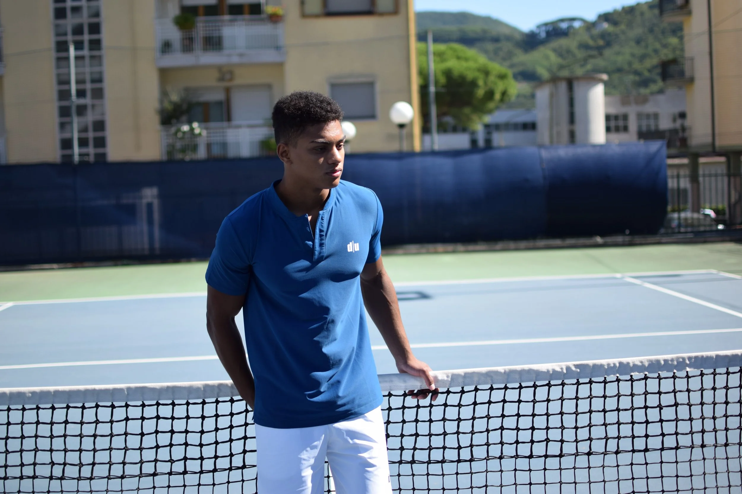 Giovane uomo triste che si appoggia alla rete di un campo da tennis all'aperto, sullo sfondo edifici e colline.