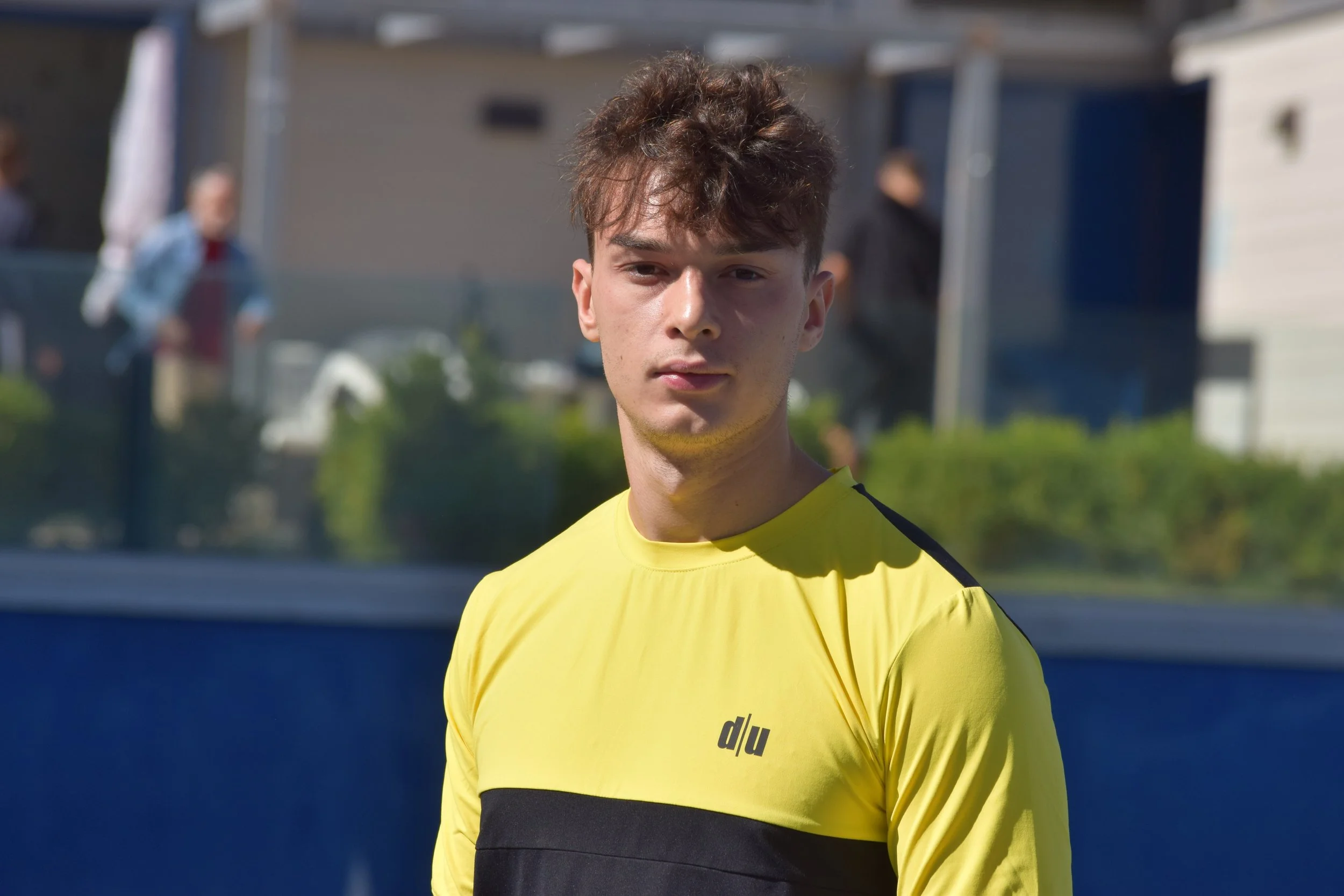 Giovane uomo con capelli ricci di fronte a un campo di sport, indossa una maglietta gialla con banda nera.