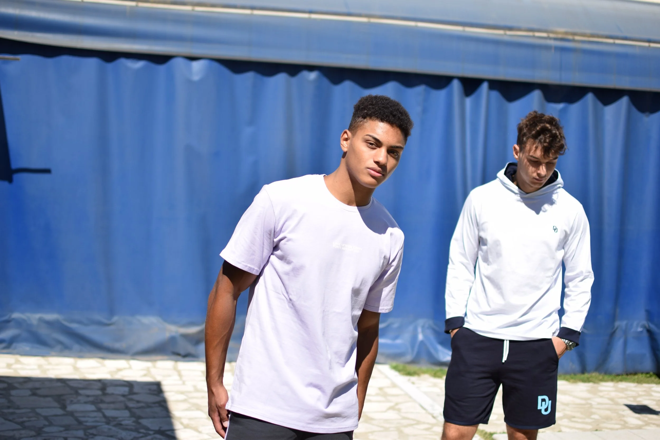 Due giovani uomini si trovano all'aperto con uno sfondo di una recinzione blu e pavimento in pietra, indossano abbigliamento sportivo casual.