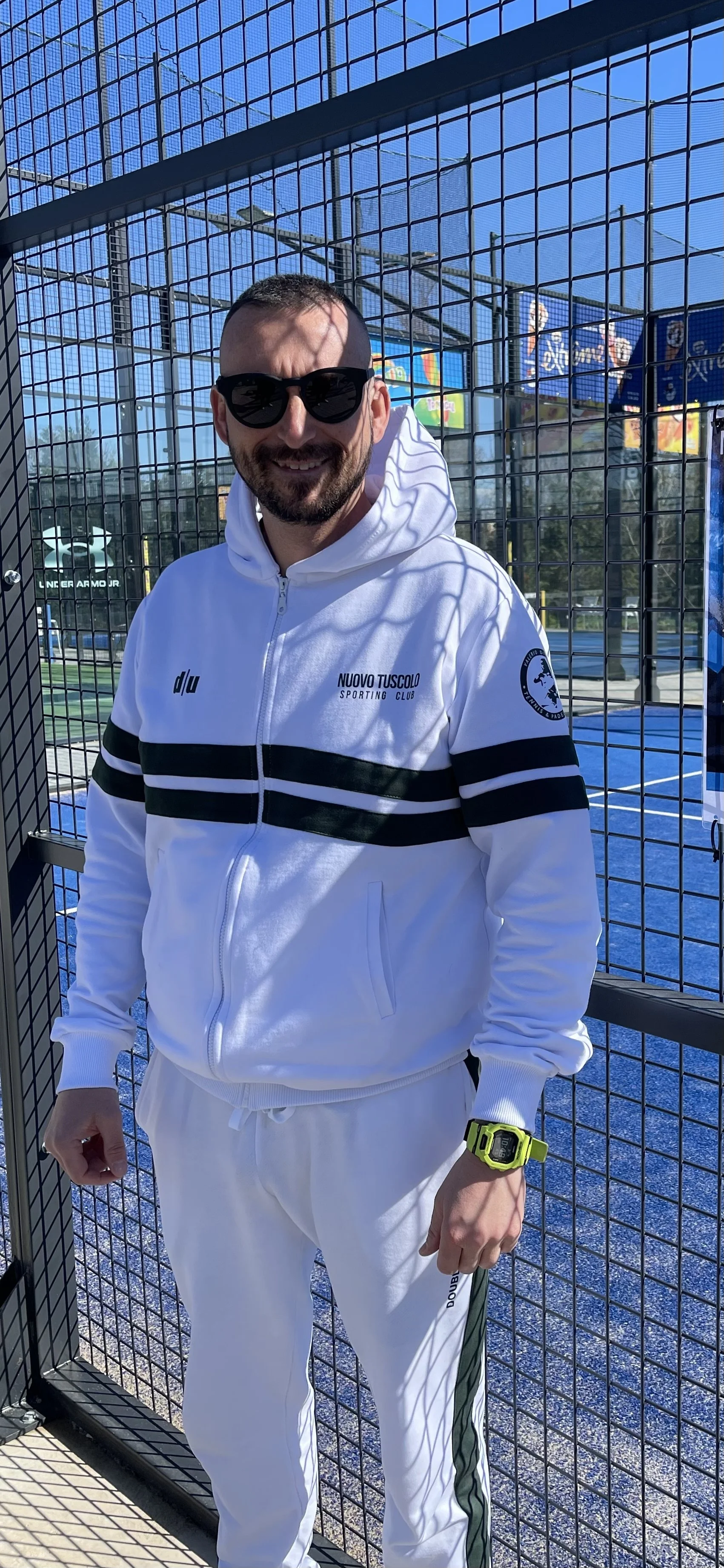 Uomo sorridente con occhiali da sole, vestito con outfit sportivo bianco con scritte, davanti a una recinzione in una area sportiva all'aperto sotto un cielo sereno.