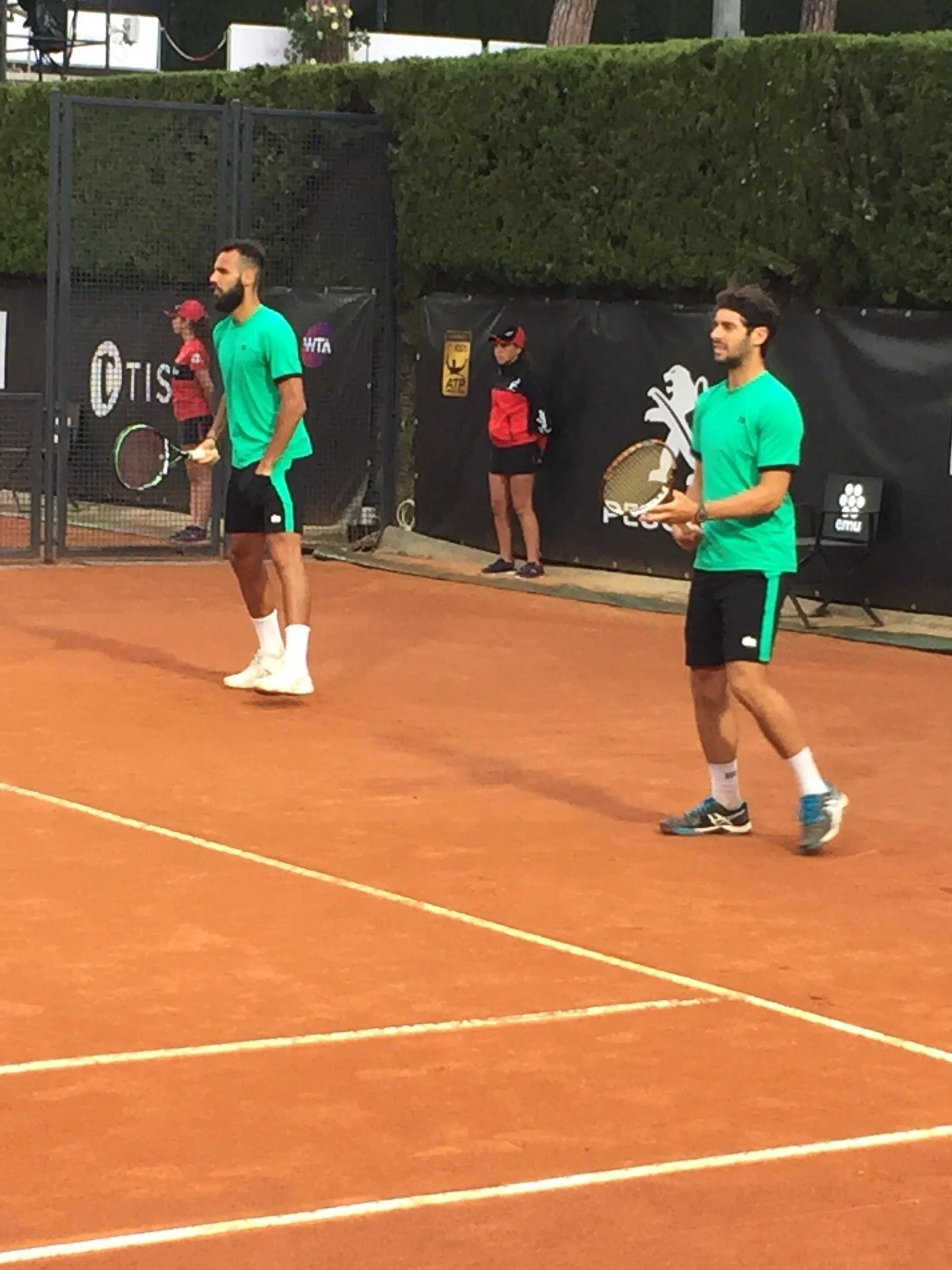 Due tennisti maschi con magliette verdi e pantaloncini neri si preparano a giocare a tennis su un campo in terra battuta, con uno sfondo di siepi e una donna con cappello e giacca rossa che osserva.