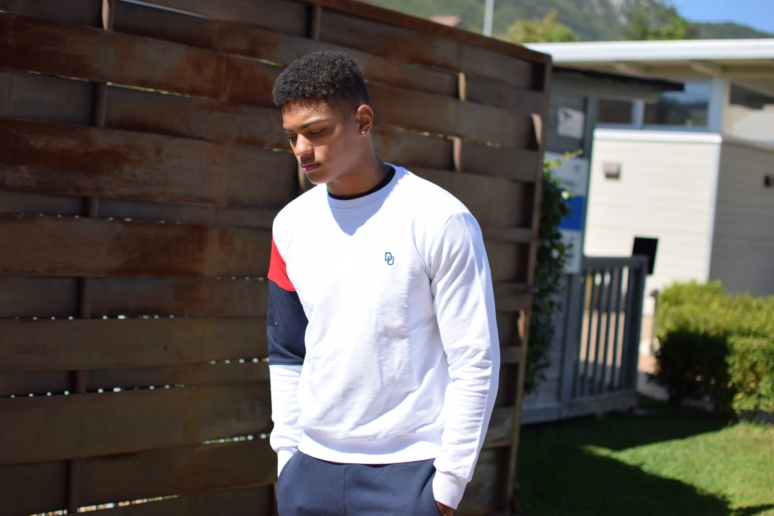 Giovane uomo con capelli corti e riccioli, indossa una felpa bianca con dettagli rossi e blu, e pantaloni scuri, sta guardando verso il basso, all'aperto.