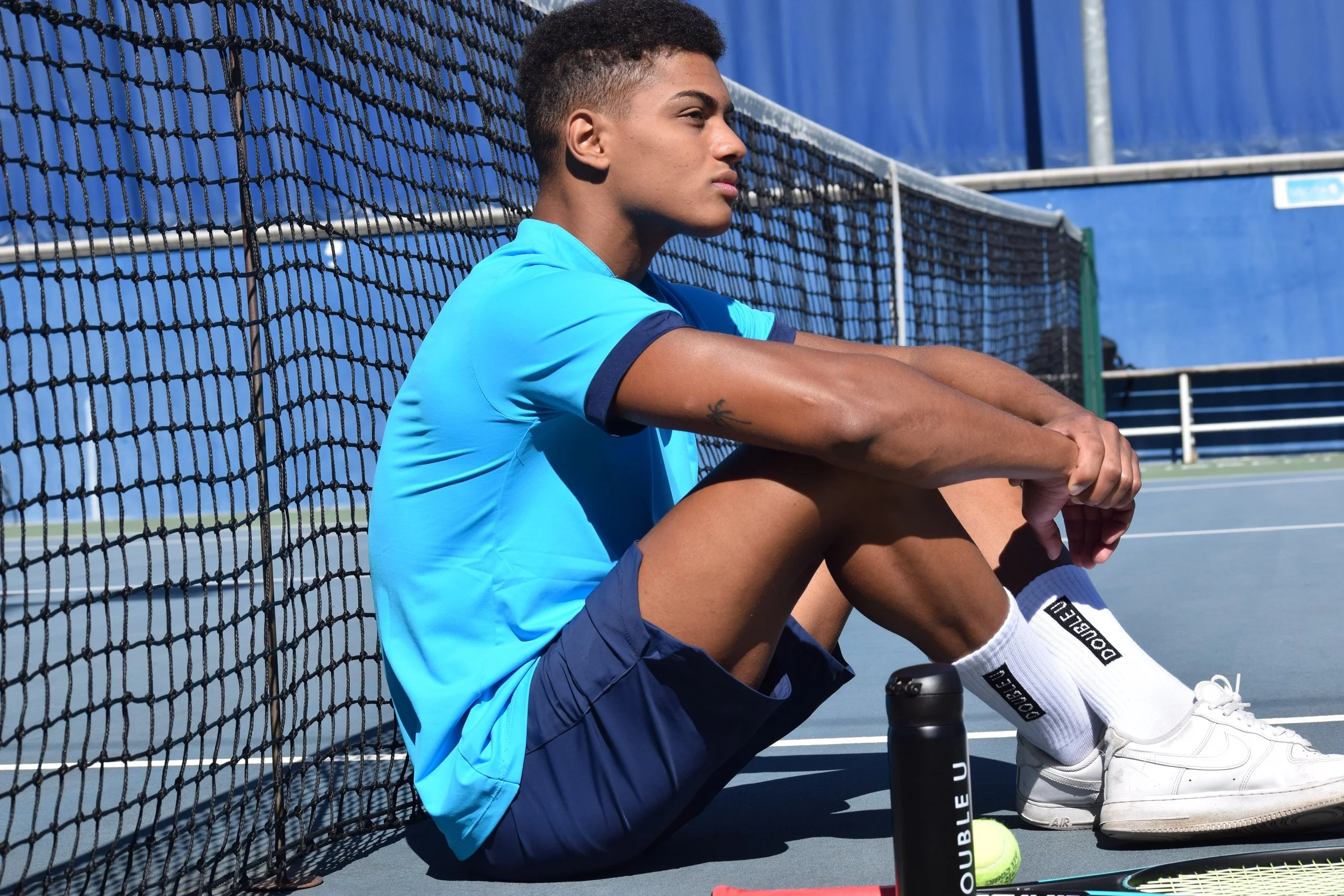 Ragazzo seduto sul campo da tennis, in abbigliamento sportivo, vicino alla rete, con attrezzatura da tennis e una palla, in un ambiente all'aperto.
