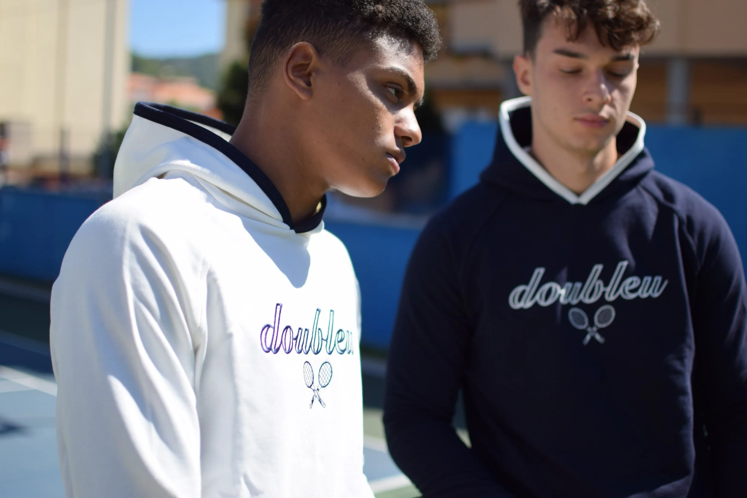 Due giovani uomini vestiti con felpe con cappuccio, uno bianco e uno nero, con il logo 'doublehnew' e due racchette da tennis sotto il testo, si trovano su un campo da tennis all'aperto