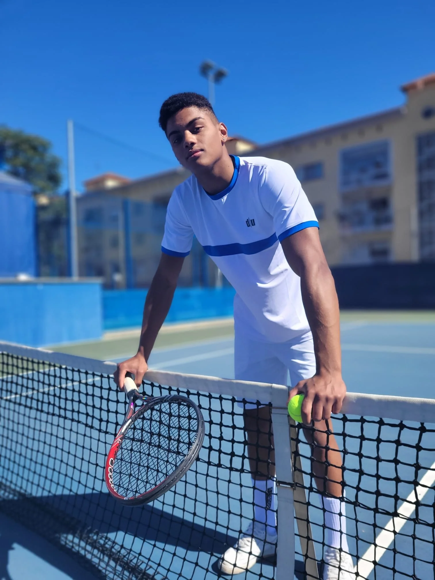 Ragazzo giovane con racchetta da tennis e palla sulla rete di un campo da tennis all'aperto in una giornata di sole.