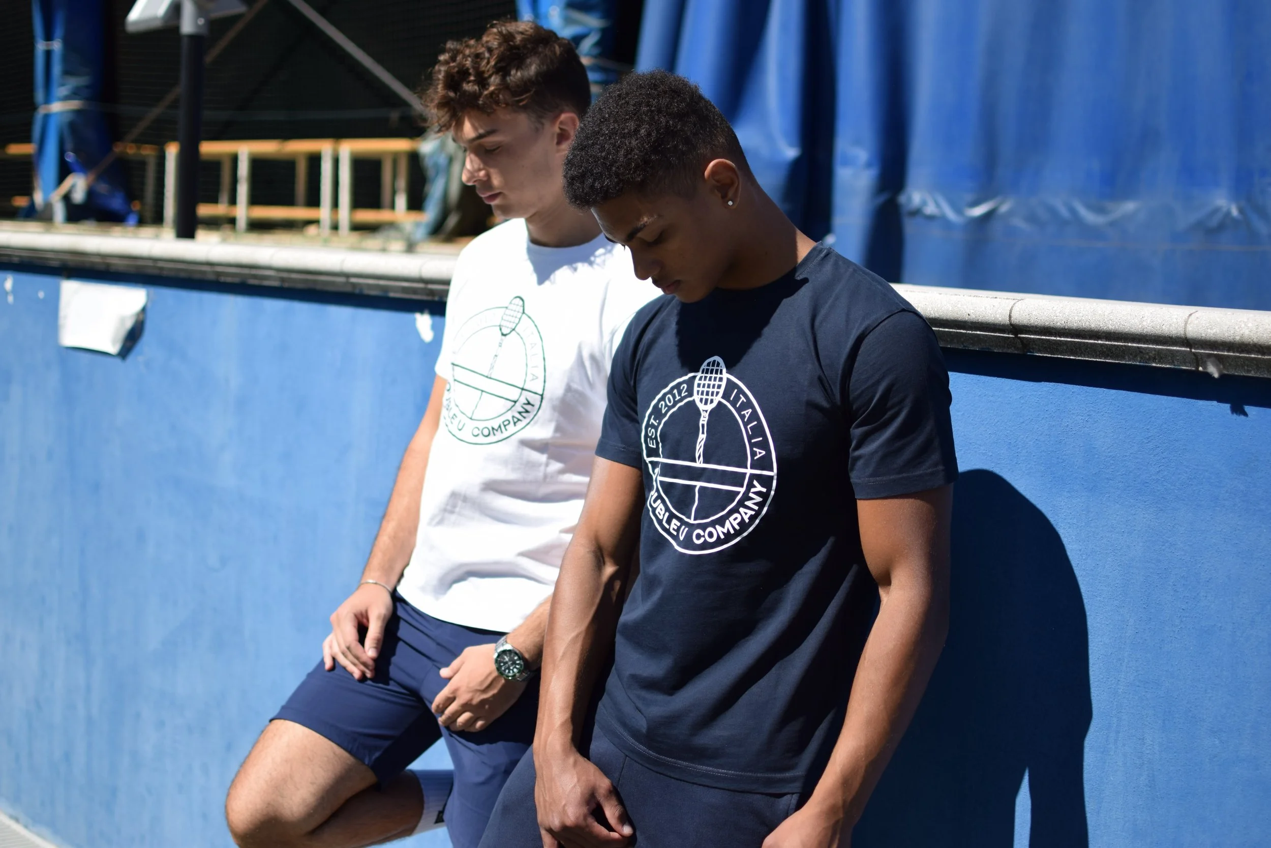 Due giovani uomini si appoggiano contro un muro blu in un giorno soleggiato, indossando magliette con il logo di una compagnia di tennis. Uno con maglietta bianca e l'altro con maglietta nera.
