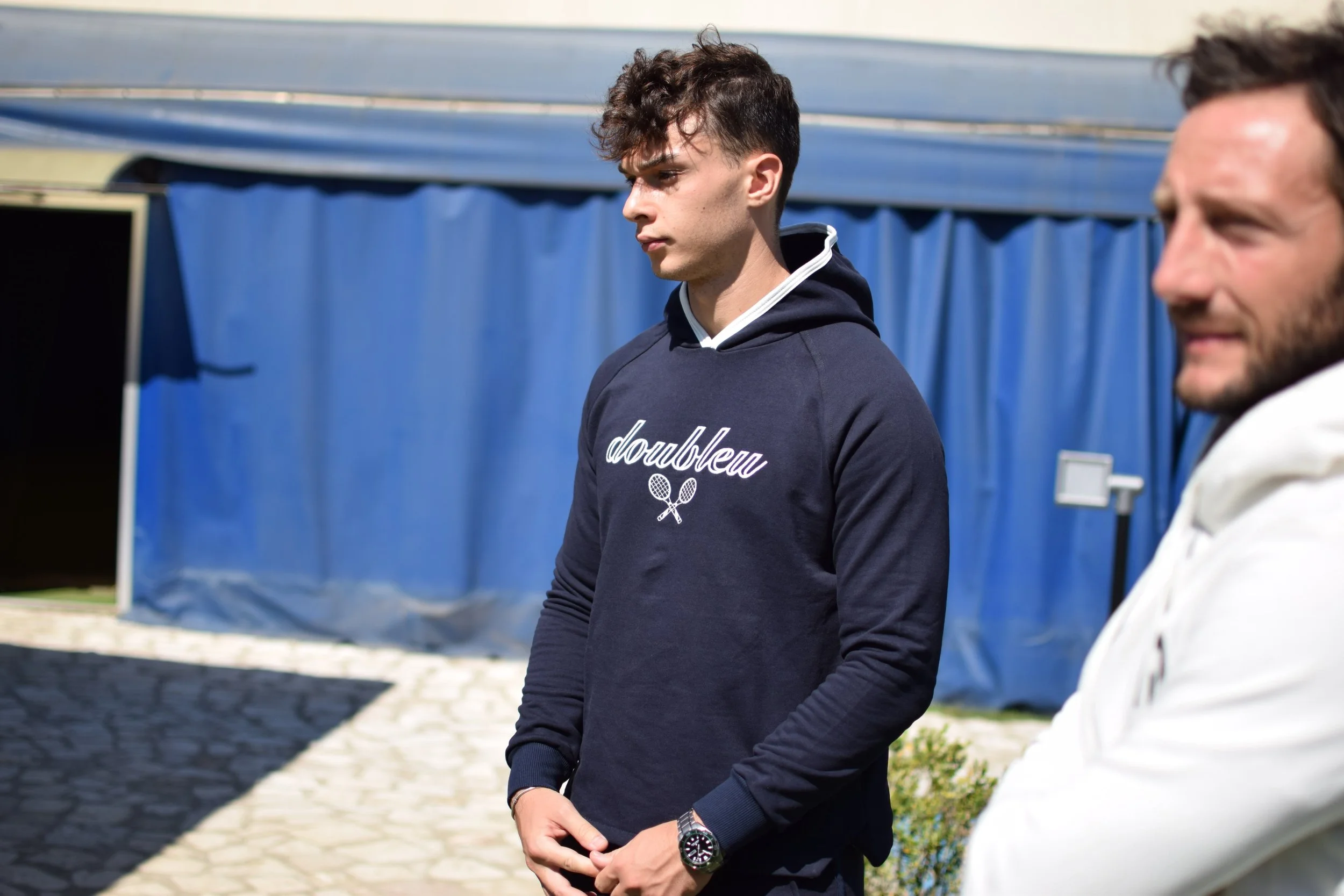 Due uomini all'aperto, uno con una felpa blu con scritta 'doubleu' e un'icona di due racchette da tennis, l'altro con una felpa bianca. C'è una struttura con tende blu sullo sfondo.