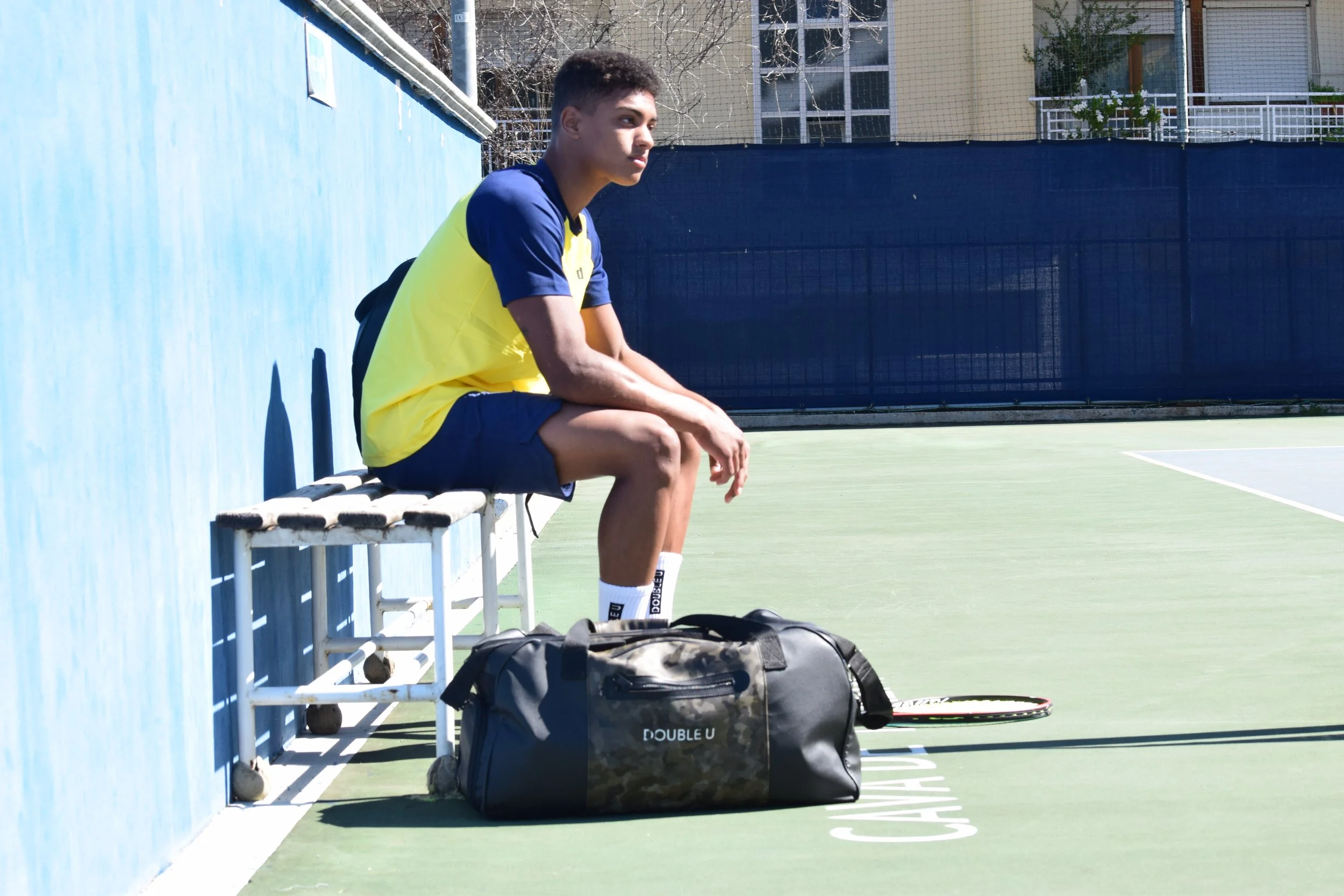 Ragazzo seduto su una panchina di un campo da tennis, con una borsa sul pavimento e una racchetta accanto