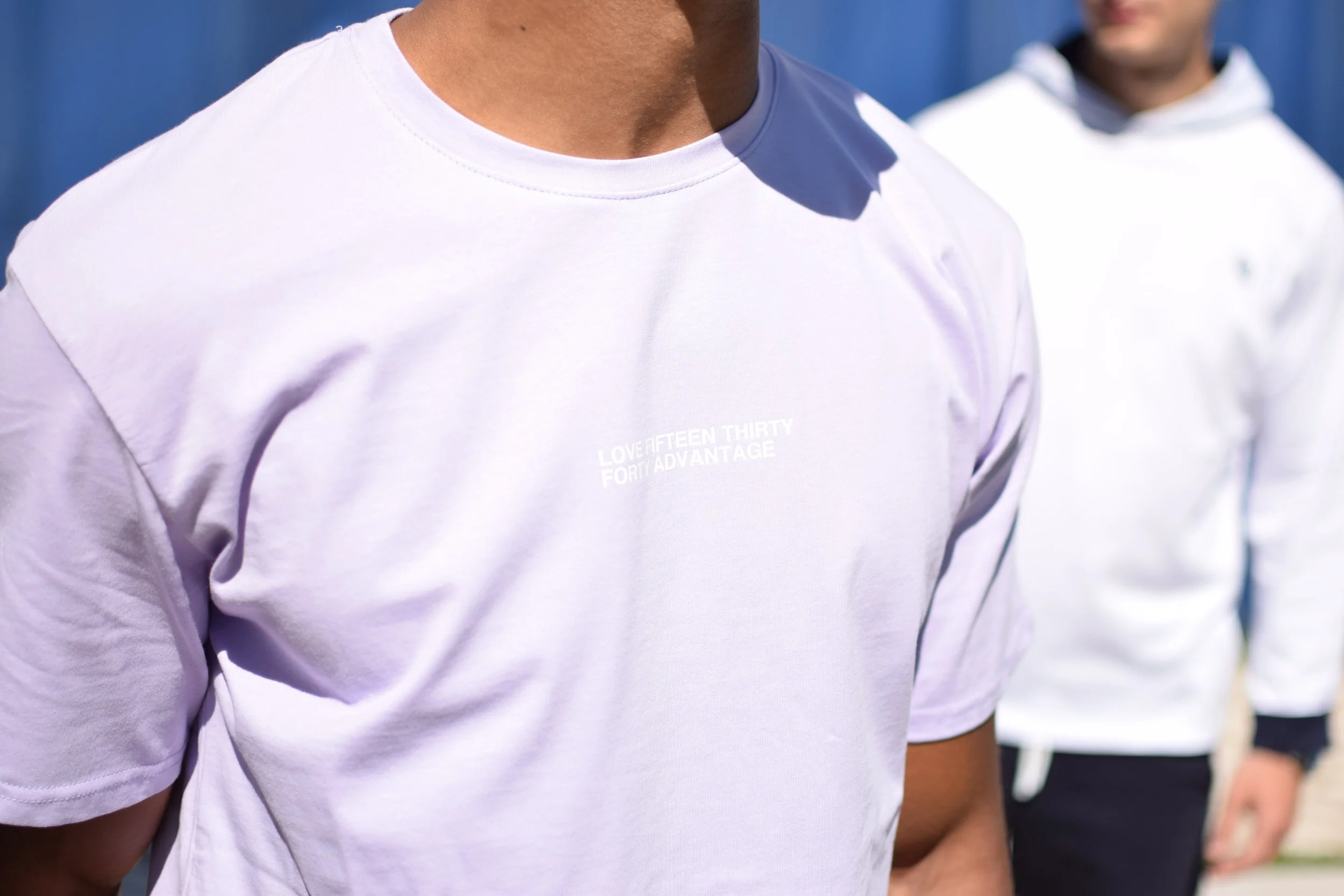 Due persone indossano magliette bianche, una in primo piano e l'altra sullo sfondo, in un ambiente esterno.