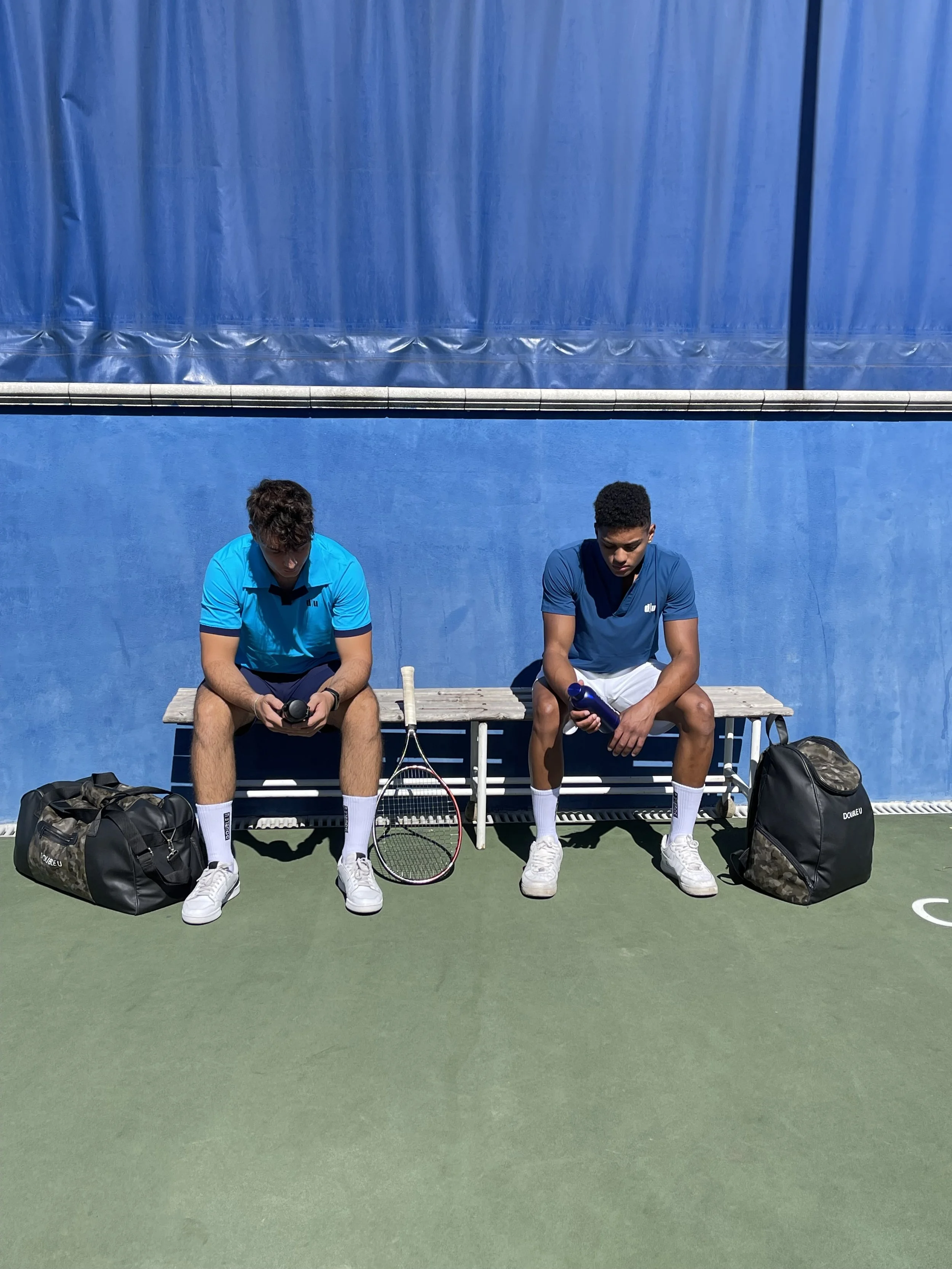 Due giovani uomini seduti su una panchina di fronte a un muro blu, con racchette da tennis e borse ai loro lati, in un campo da tennis all'aperto.