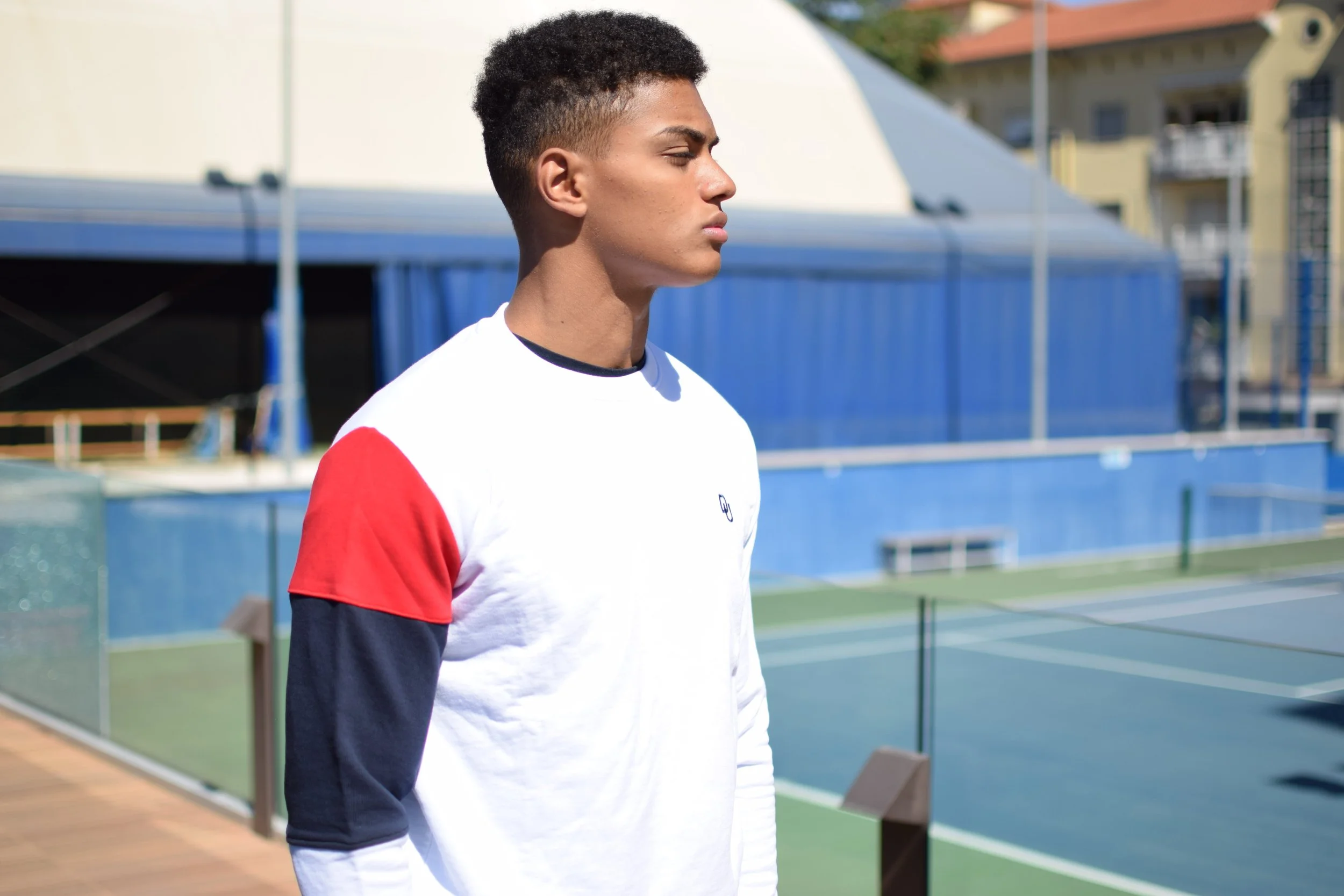 Ragazzo con maglietta bianca con maniche a colori rosso e nero, che si trova vicino a un campo da tennis all'aperto