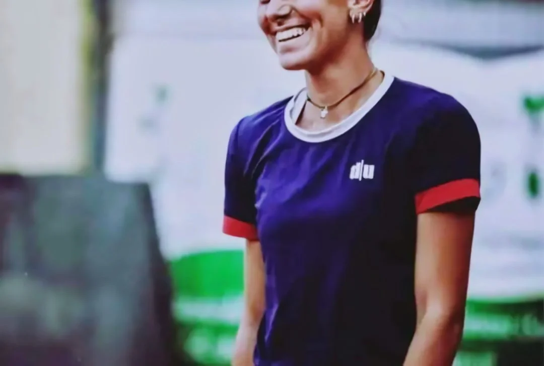 ritratto di una donna sorridente con braccio nudo, indossa una maglietta blu con dettagli rossi e accessori come orecchini e una collana