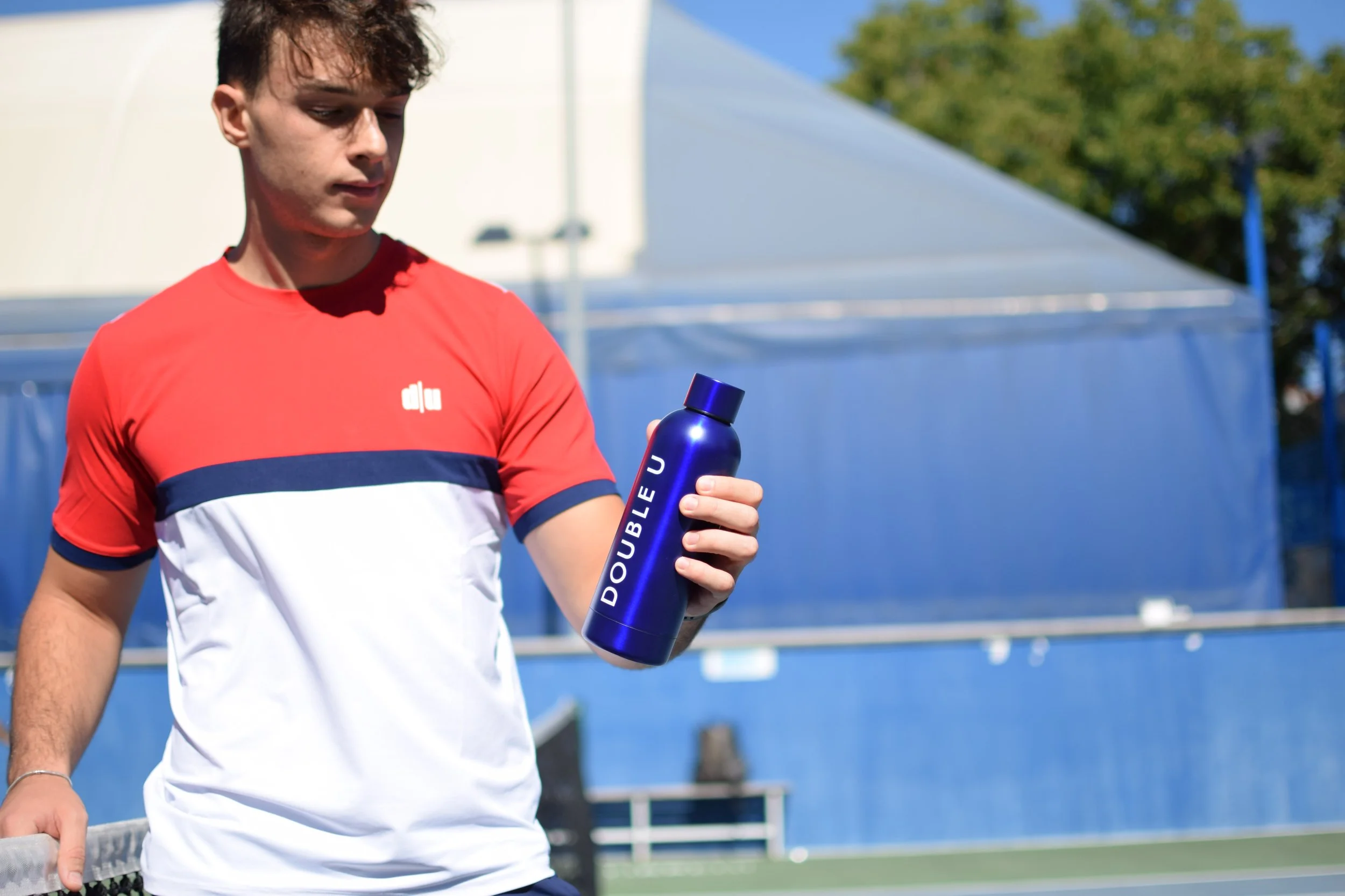 Giovane tennisman tiene una bottiglia blu con la scritta DOUBLEU, indossa una maglietta rossa e bianca, sul campo di tennis con una struttura blu sullo sfondo.