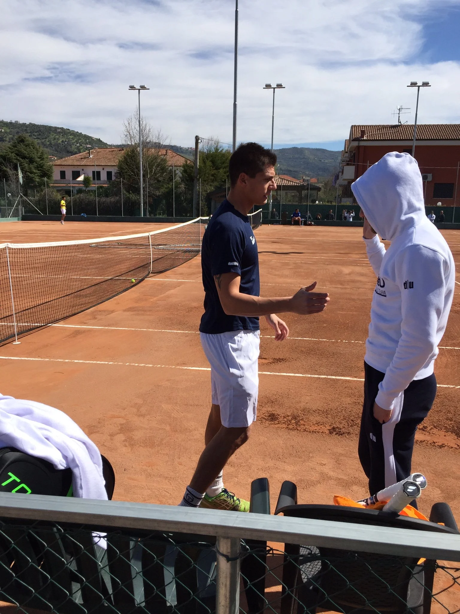 Due giocatori di tennis discutono sulla rete di un campo da tennis in campo all'aperto con montagne sullo sfondo, uno indossa una t-shirt blu e pantaloncini bianchi, l'altro una felpa bianca con cappuccio.