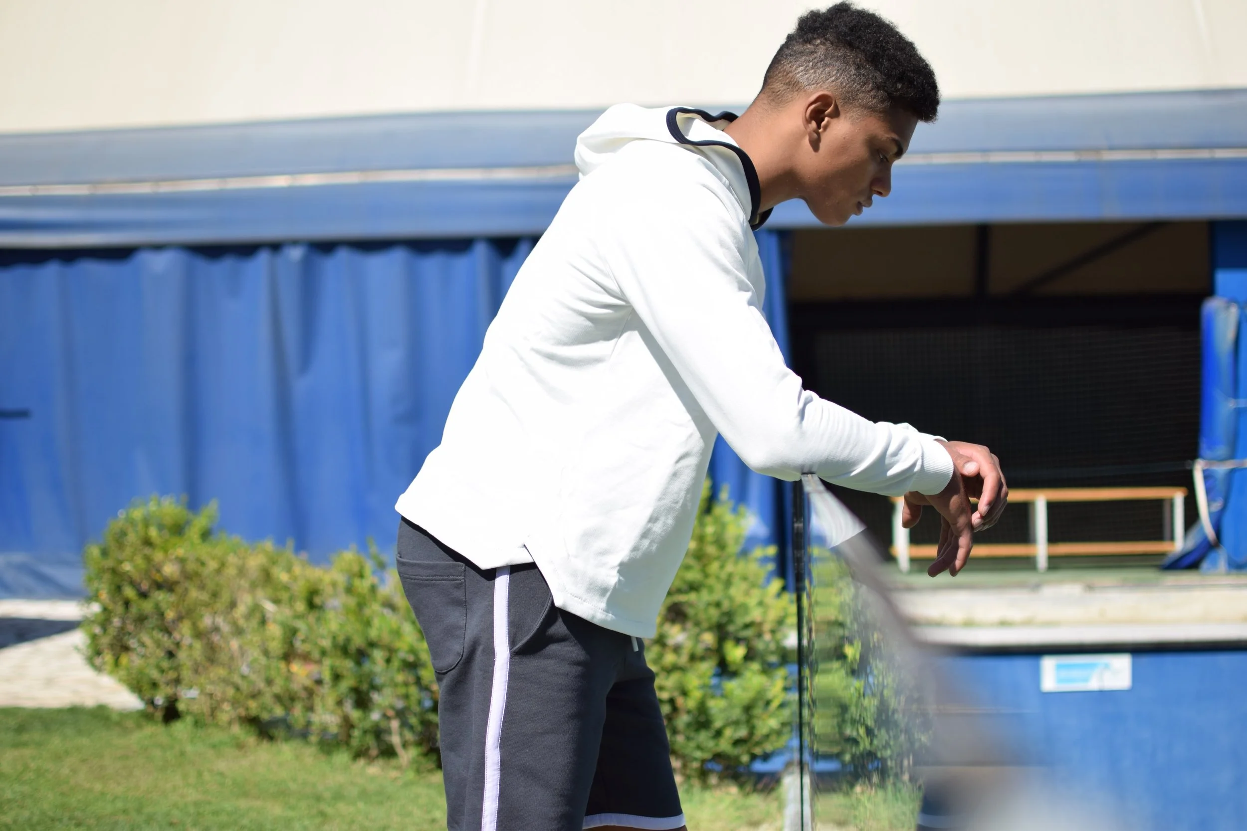 Un giovane uomo in piedi in un campo di tennis, guarda un orologio al polso, indossa una felpa bianca e pantaloni sportivi grigi con strisce bianche. Sullo sfondo, tende blu e arbusti verdi.