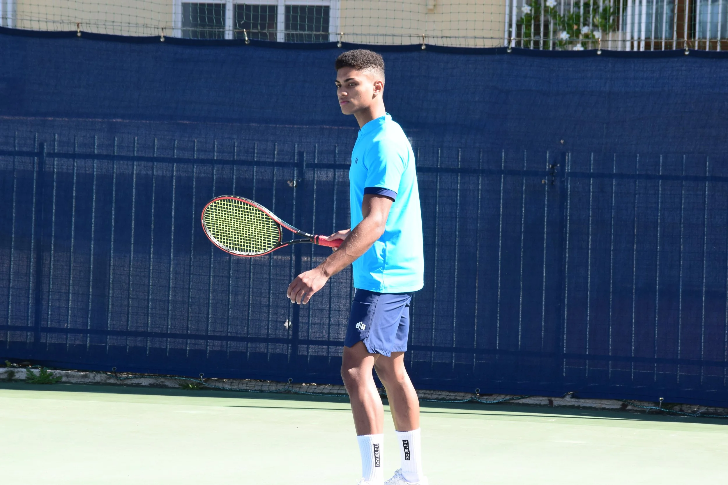 Giovane uomo che gioca a tennis su un campo all'aperto, indossa una maglietta azzurra, pantaloncini blu e calze bianche.