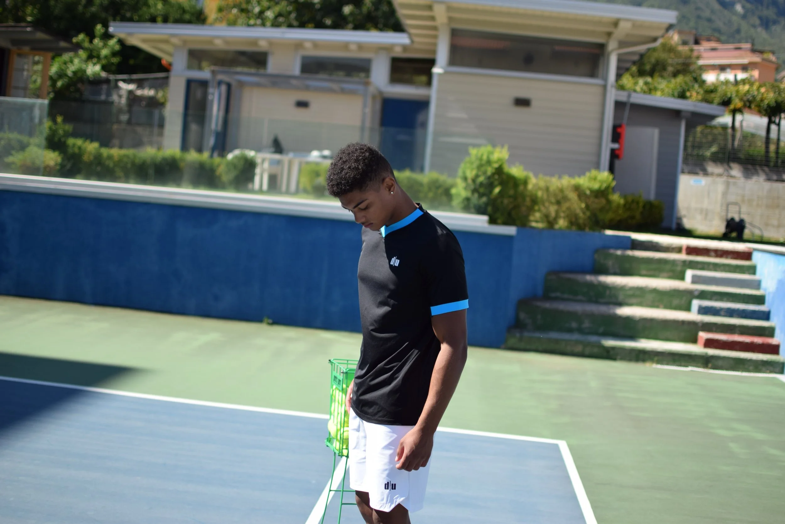 Un giovane uomo con maglietta nera e pantaloncini bianchi su un campo da tennis, con le mani ripiegate davanti, guarda verso il basso, sullo sfondo una piscina e alcune scale.