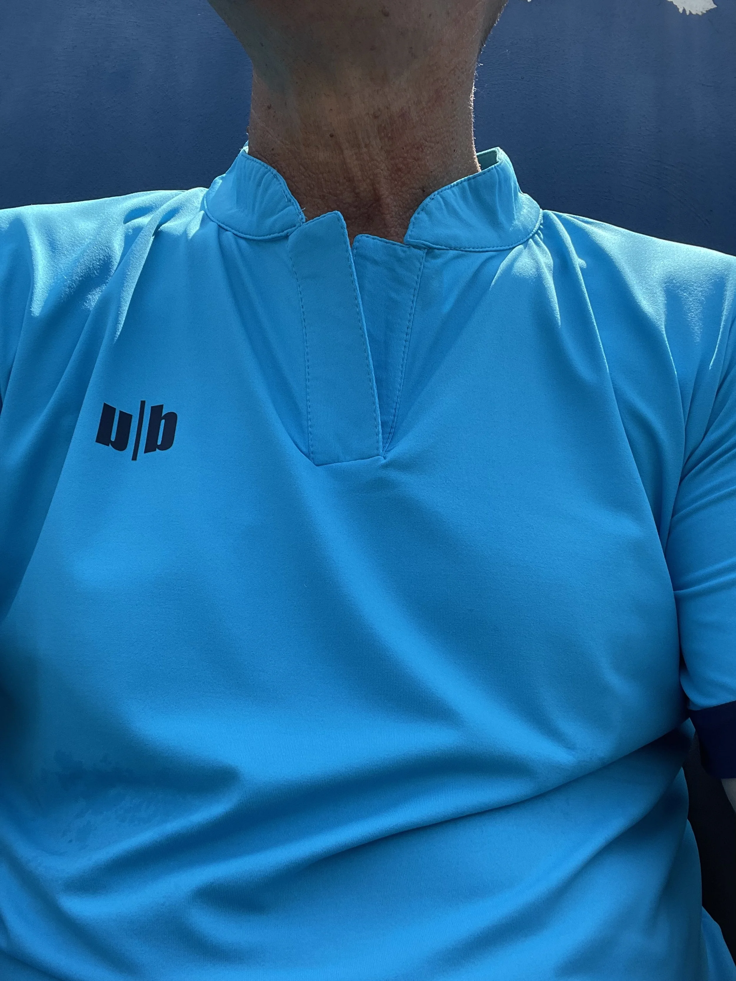 Parte superiore di una persona con una maglietta sportiva azzurra, scollo a V e logo blu sulla spalla sinistra.