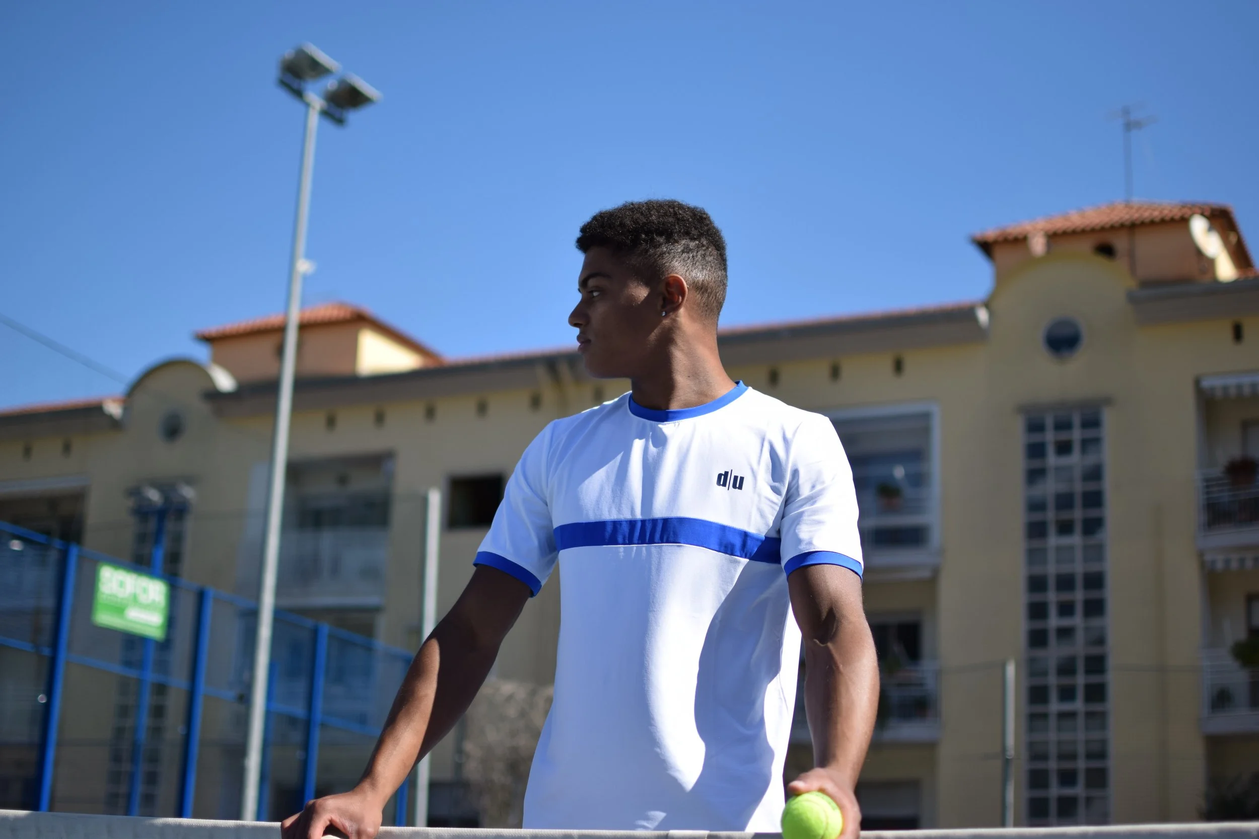 Giocatore di tennis giovane con maglietta bianca e dettagli blu, che tiene una pallina da tennis, in un campo all'aperto con edificio e lampione sullo sfondo, sotto un cielo blu.