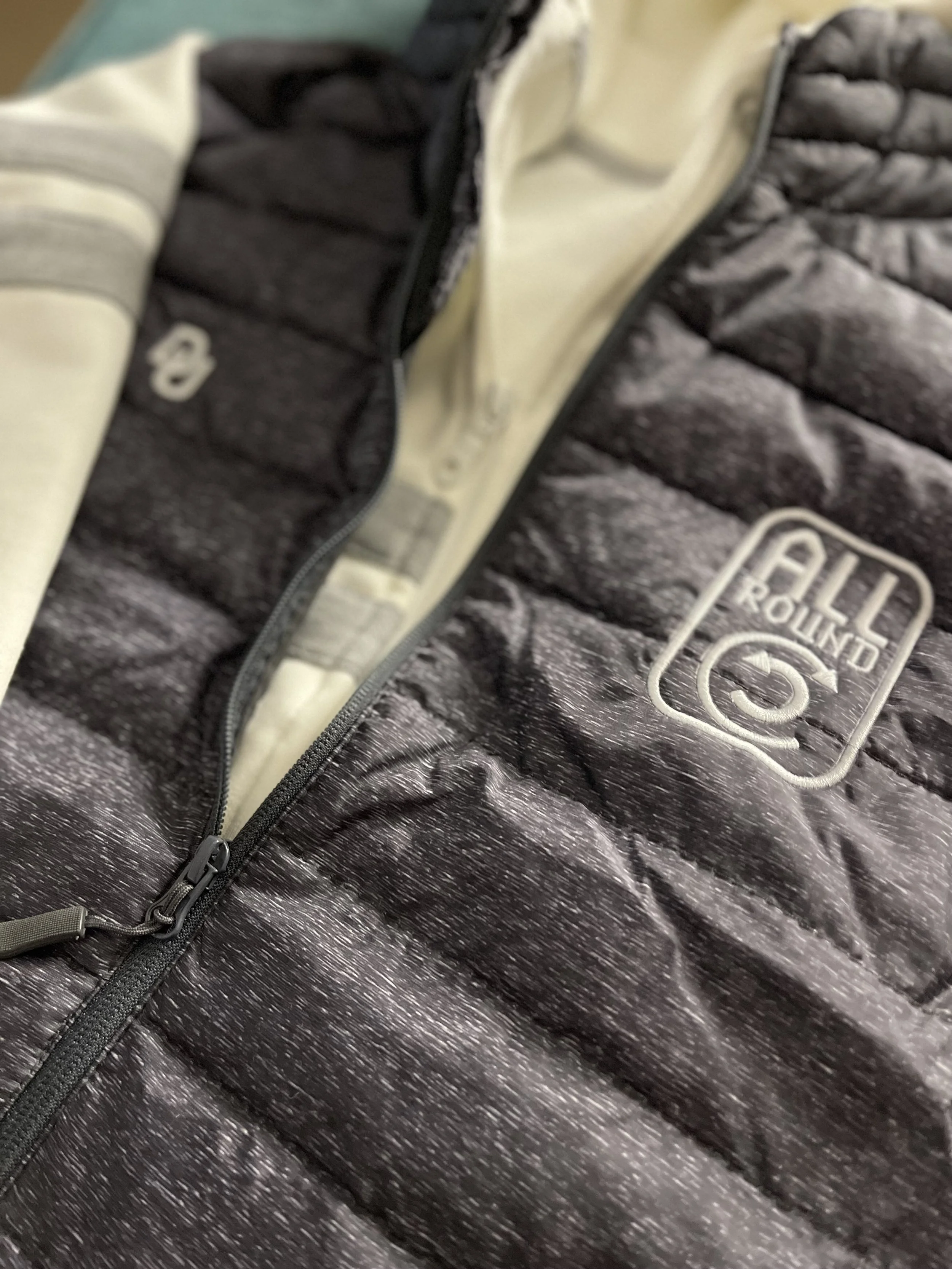 Dettaglio di una giacca imbottita con zip, logo e patch sulla parte anteriore, colore grigio con lavorazione a righe.