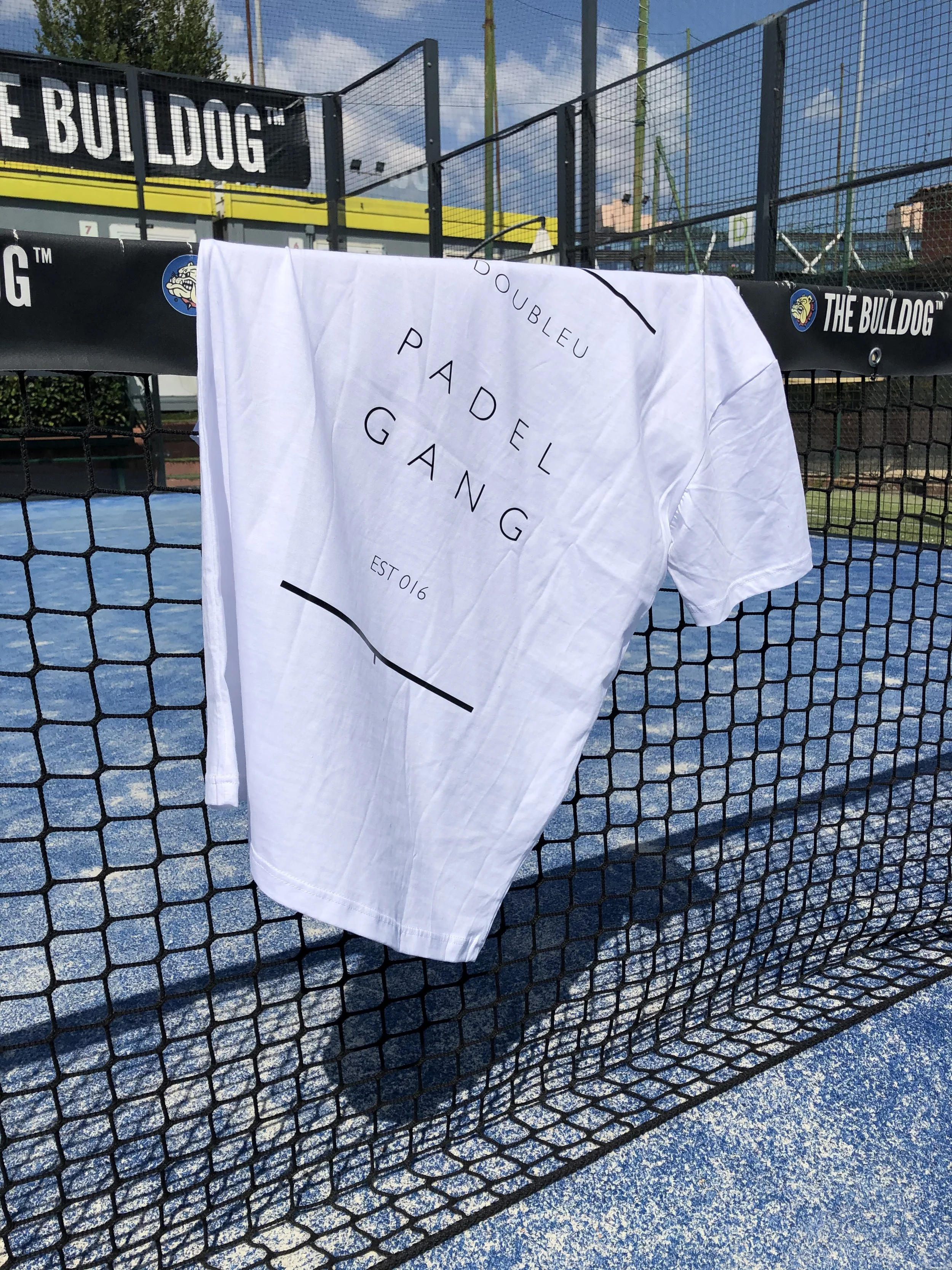 Asciugamano bianco appeso sulla rete di una campo da padel, con scritte nere: 'Doubleu', 'Padel Gang', 'Est 016', e linee decorative