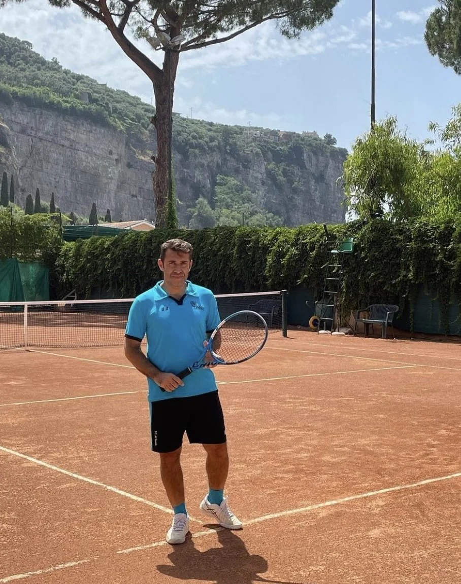 Uomo con maglietta azzurra, pantaloncini neri e scarpe bianche che tiene una racchetta da tennis su un campo di tennis in terracotta, con alberi e montagne sullo sfondo.