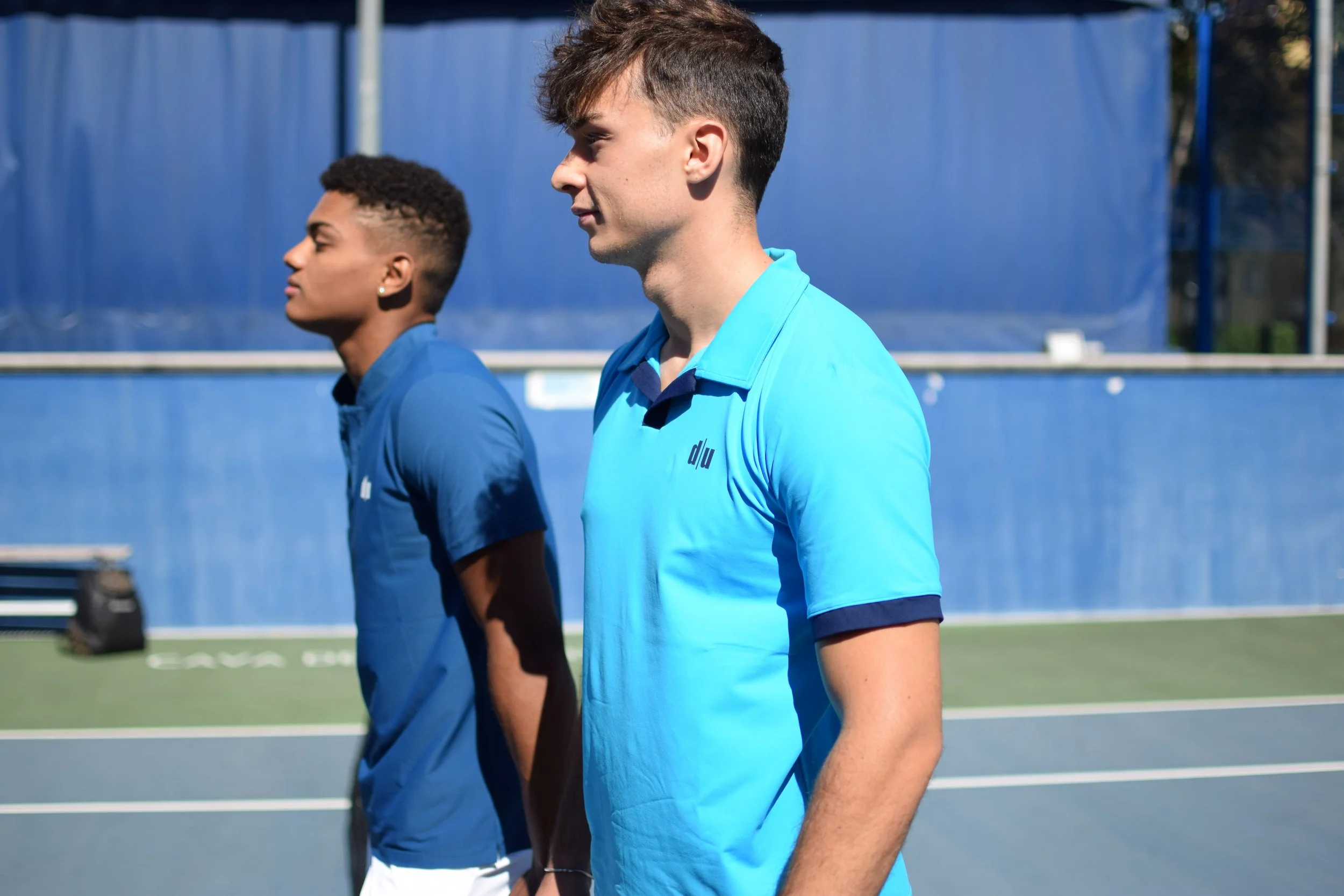 Due giovani uomini in maglietta azzurra che si allenano su un campo da tennis all'aperto.