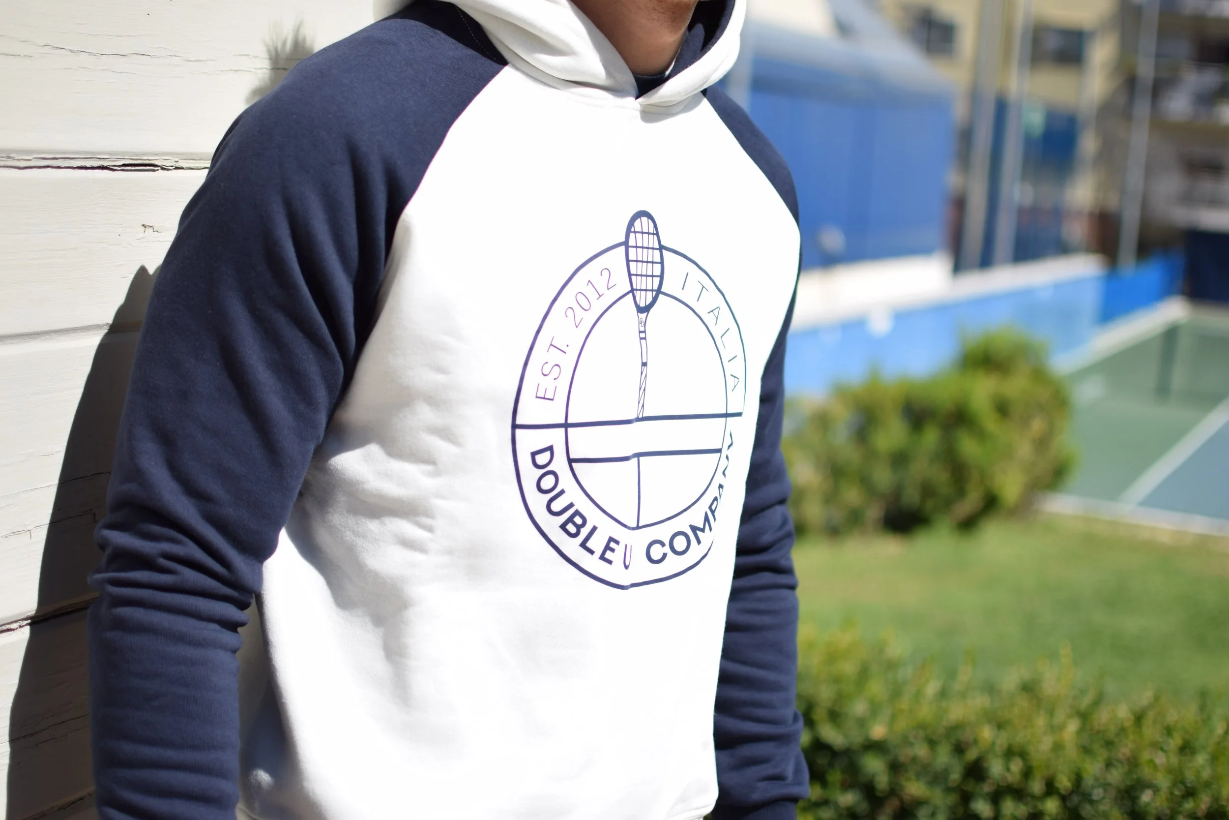 Persona che indossa una felpa bianca e blu con logo di tennis, sul retro di una rete da tennis e un campo di gioco all'aperto.