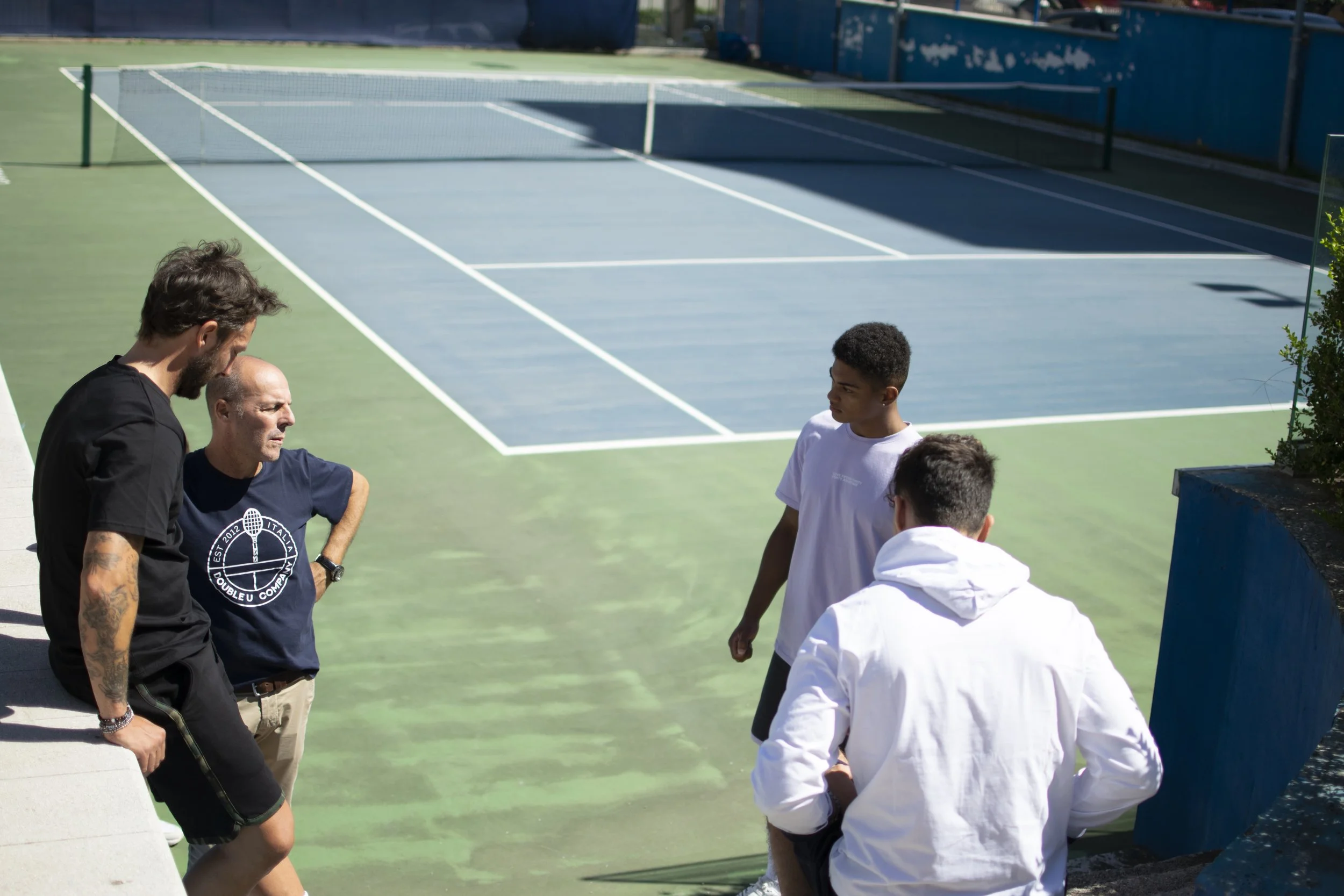 Quattro persone, tra cui due giovani e due adulti, si trovano vicino a un campo da tennis all'aperto, discutendo o ascoltando con attenzione. È una giornata luminosa e soleggiata.