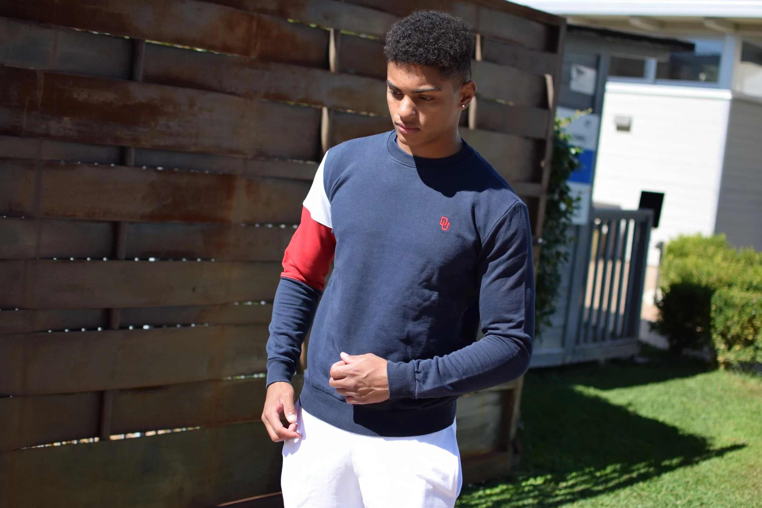 Giovane ragazzo in abbigliamento sportivo, con felpa blu con dettagli rossi e bianchi, e pantaloni bianchi, che si trova in giardino con recinzione in legno e vegetazione verzi.