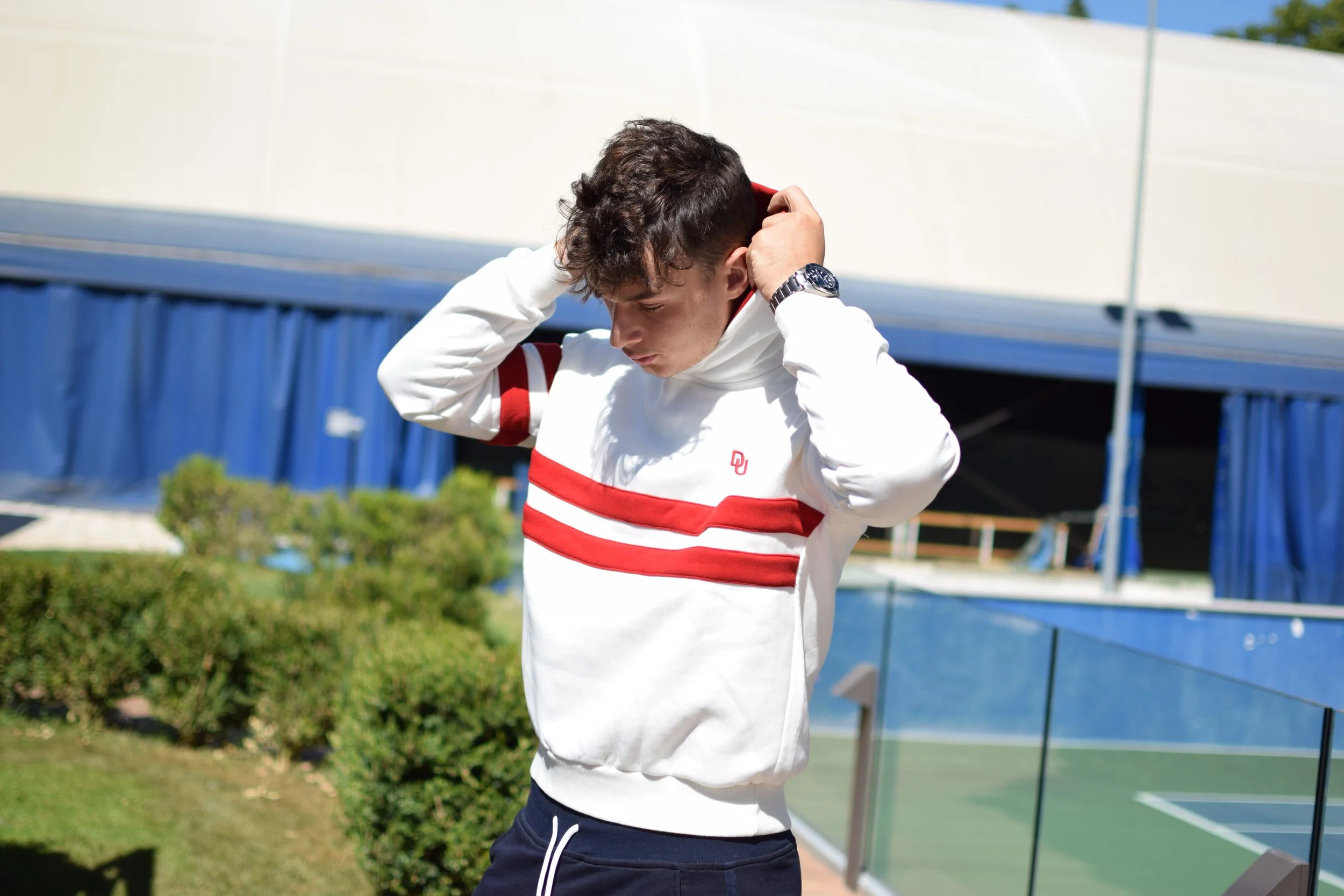 Giovane uomo con cappuccio bianco con strisce rosse, in piedi in un'area all'aperto vicino a un campo sportivo, che si sistema il cappuccio.