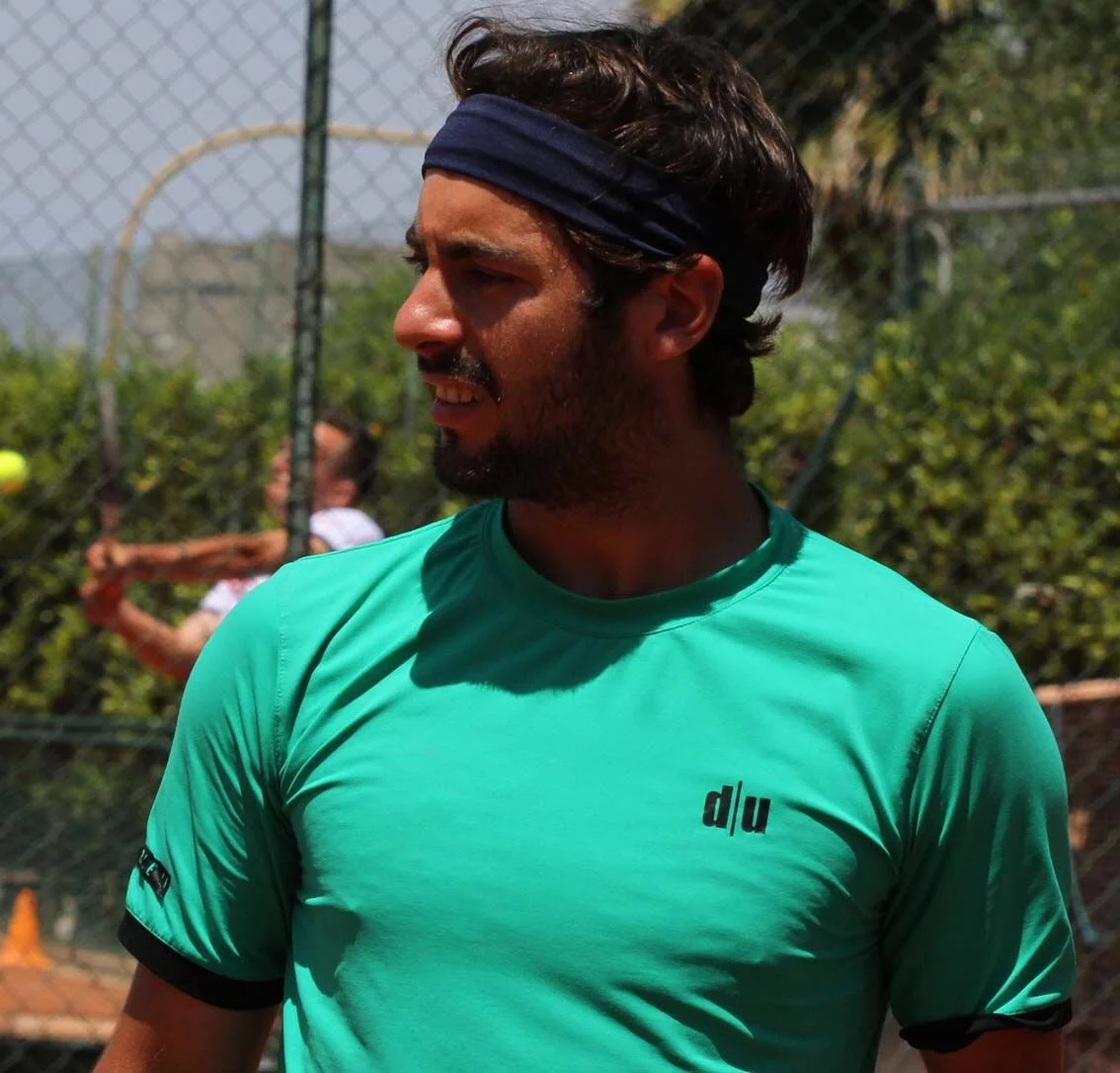 Uomo con cappello nero e maglietta verde che gioca a tennis all'aperto con rete e altre persone sullo sfondo.