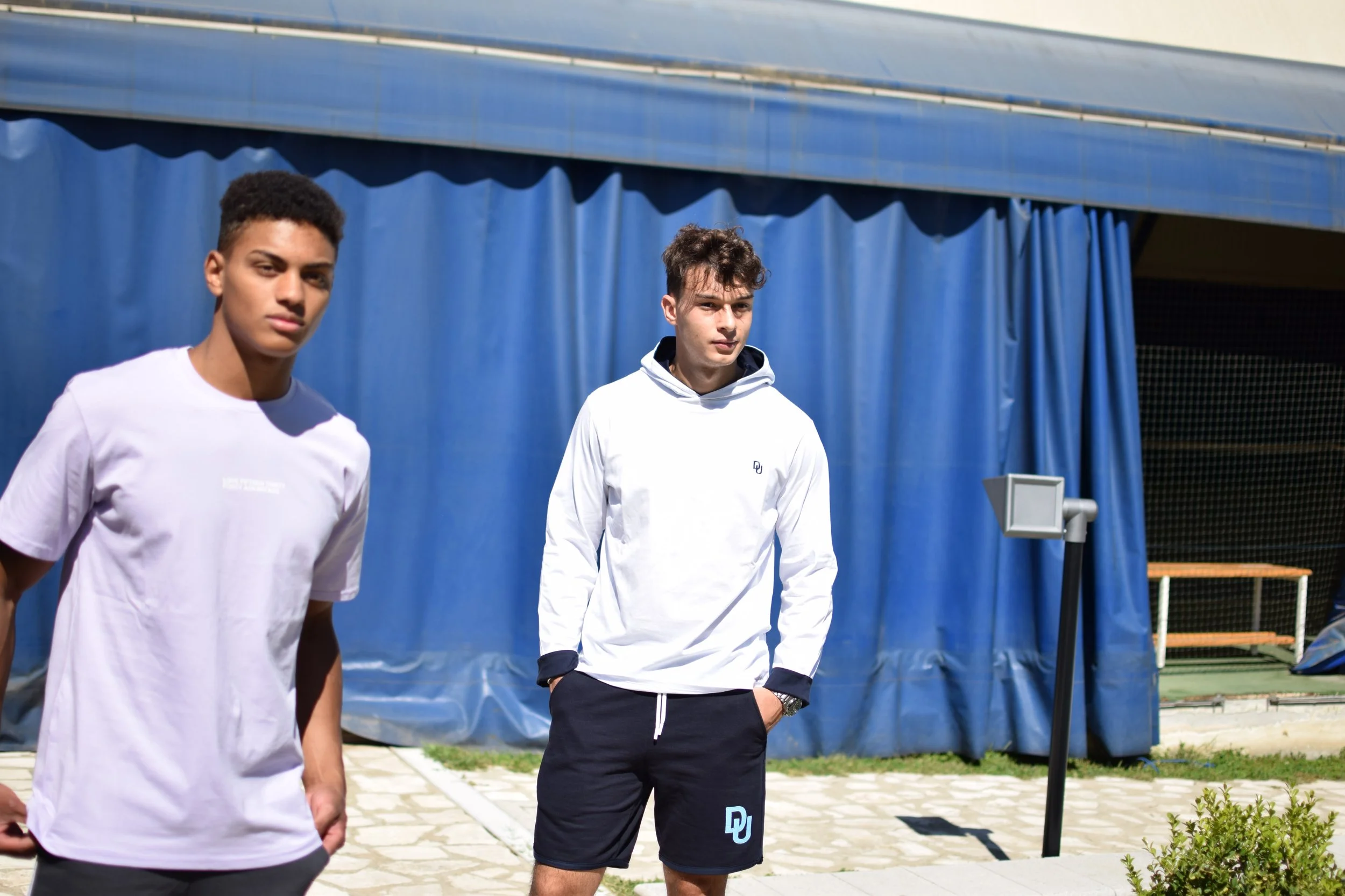 Due giovani uomini in abbigliamento sportivo si trovano all'aperto davanti a una struttura coperta con tende blu. Cassandra ergono in posizione rilassata, uno guarda direttamente la fotocamera, l'altro si distingue per i pantaloncini corti e una magl