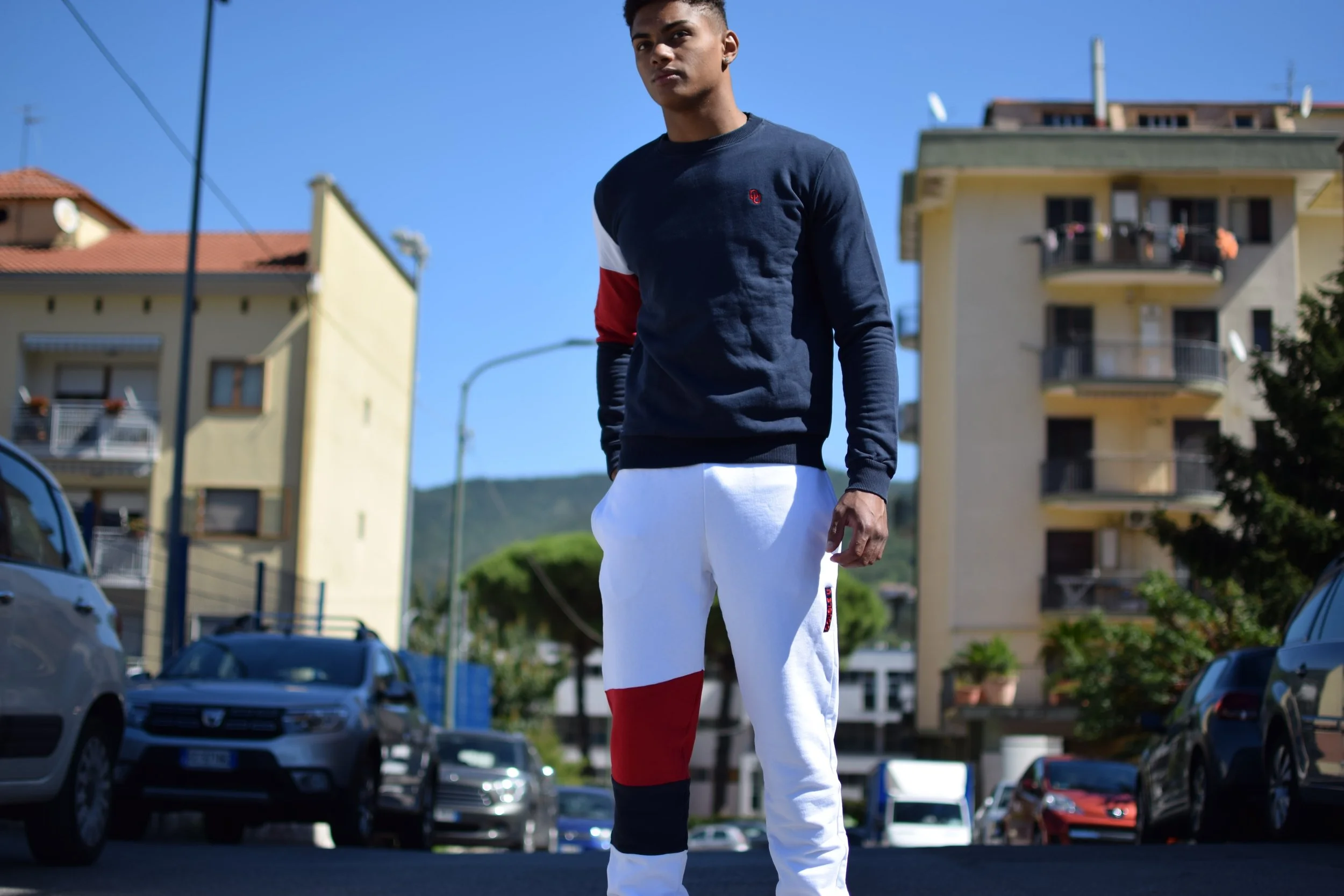 Un giovane uomo sta in piedi in una strada di città, indossa una felpa blu con dettagli rossi e bianchi e pantaloni bianchi con dettagli rossi e neri. Sullo sfondo ci sono edifici e auto parcheggiate.