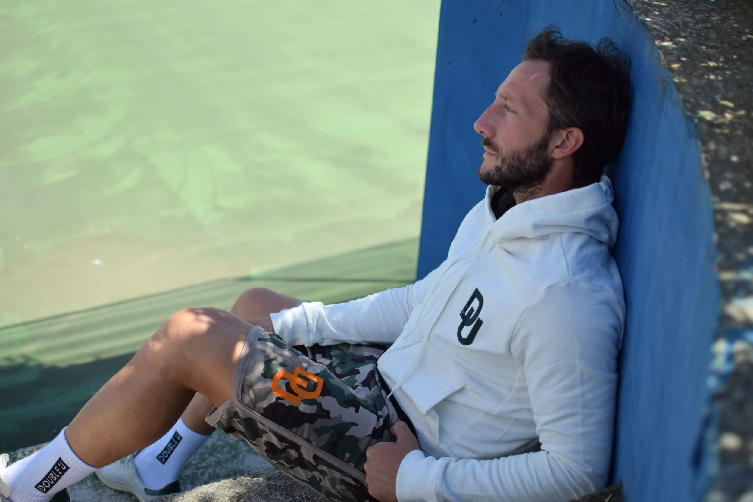 Uomo sdraiato contro una parete blu vicino ad un lago, vestito con abbigliamento sportivo. 