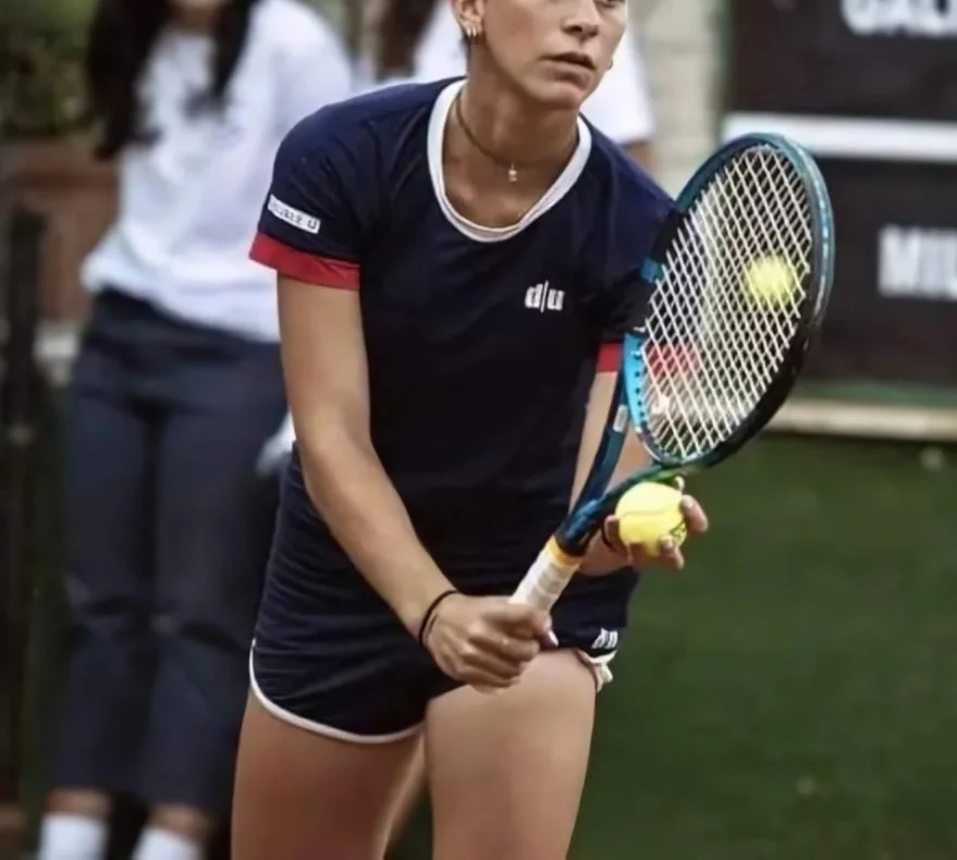 Una donna che gioca a tennis, tiene in mano una racchetta e una palla da tennis, indossa un vestito sportivo nero con dettagli rossi e bianchi.