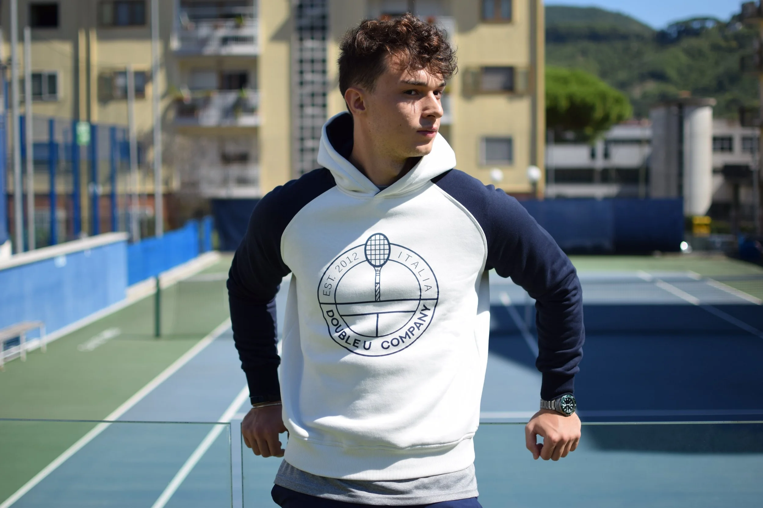 Un giovane uomo con una felpa bianca e calzoni blu si allena su un campo da tennis all'aperto, con edifici e colline sullo sfondo.