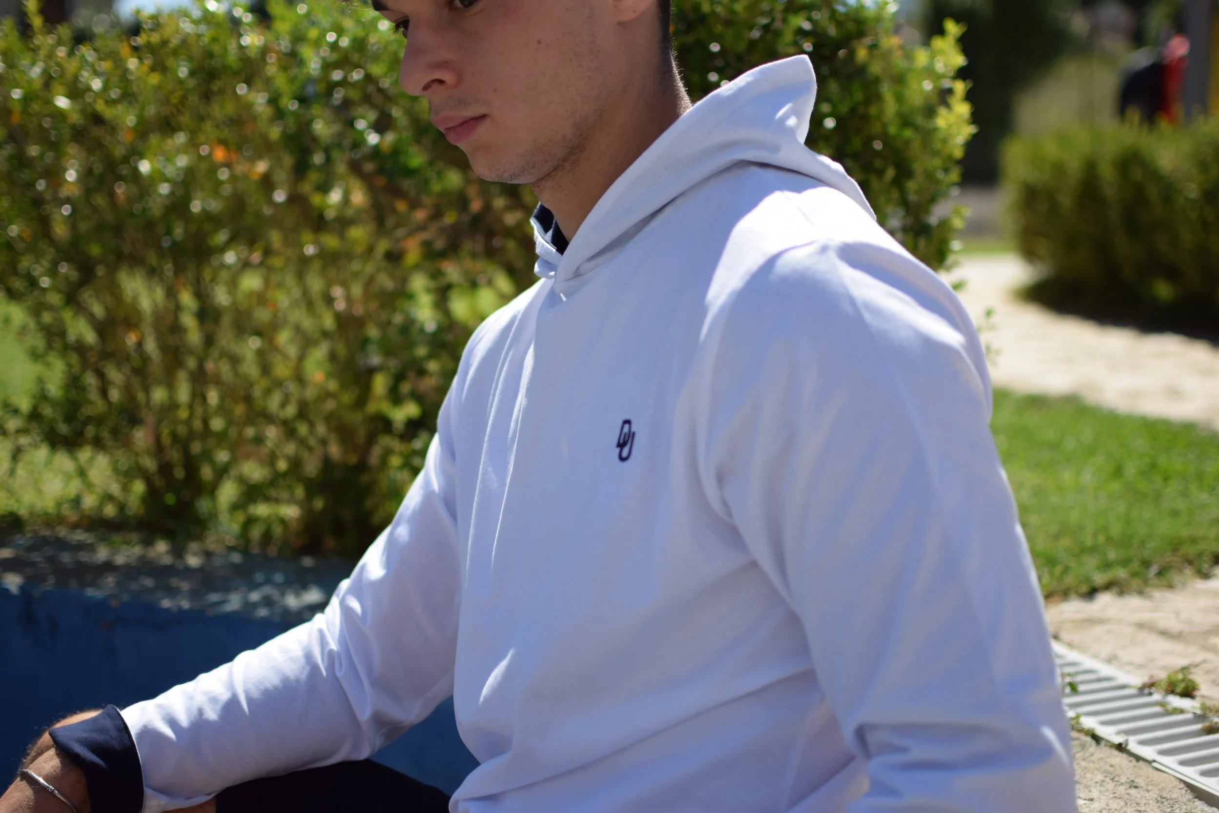 Un giovane uomo seduto all'aperto, indossa una felpa bianca con un logo piccolo e scuro, il contesto è un'area verde con cespugli e un sentiero.