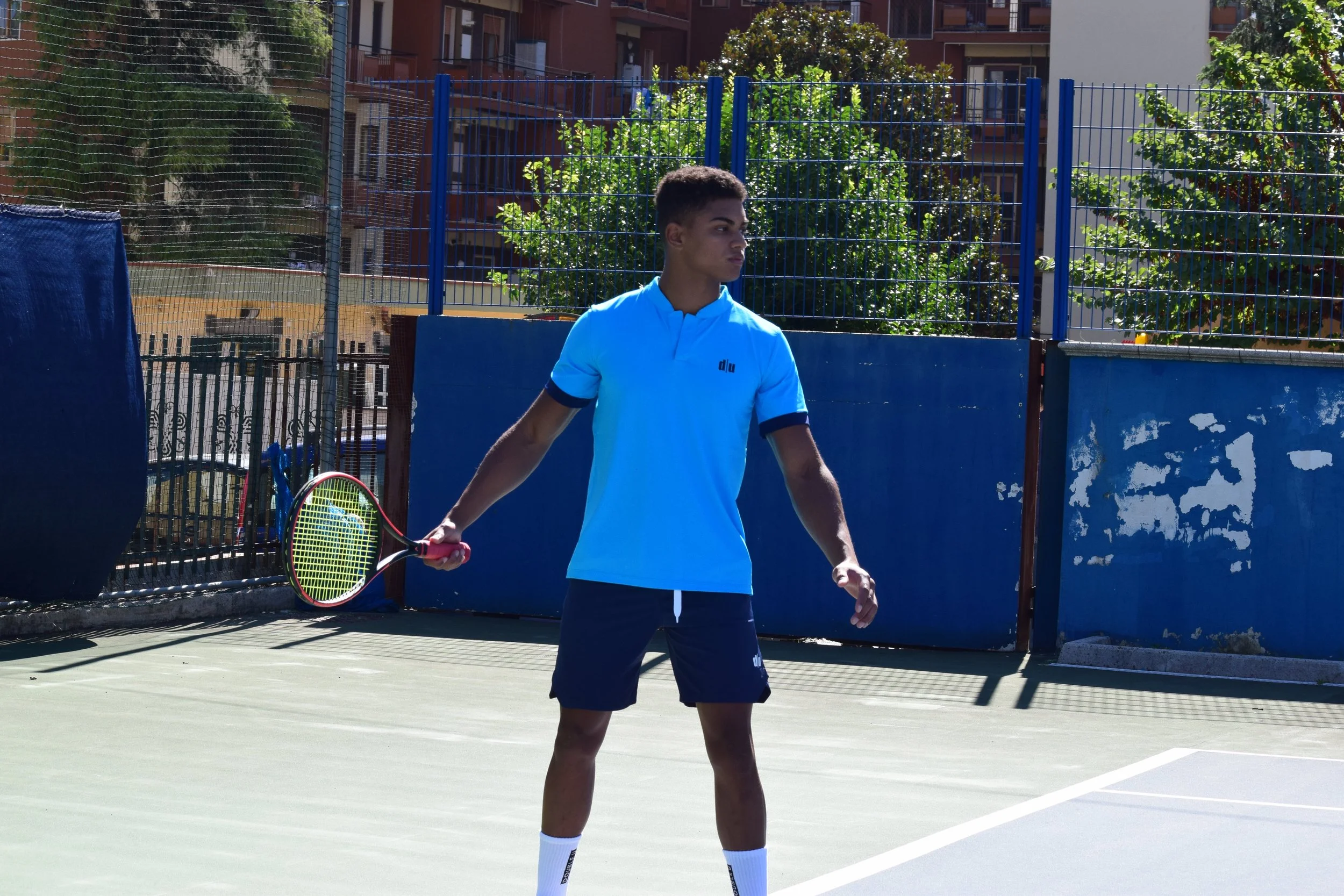 Un giovane uomo che gioca a tennis su un campo all'aperto, con una racchetta in mano, indossando una maglietta azzurra e pantaloncini neri.